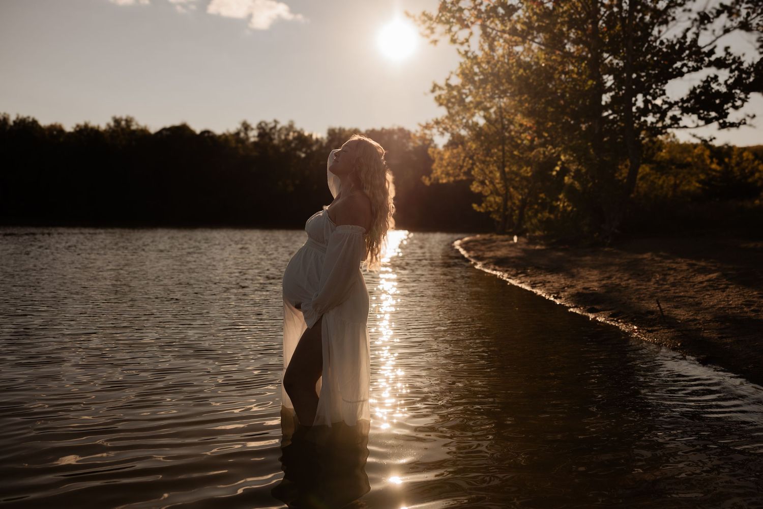 Figure silhouetted in glowing sunlight stands in lake water along wooded shoreline during golden hour sunset.