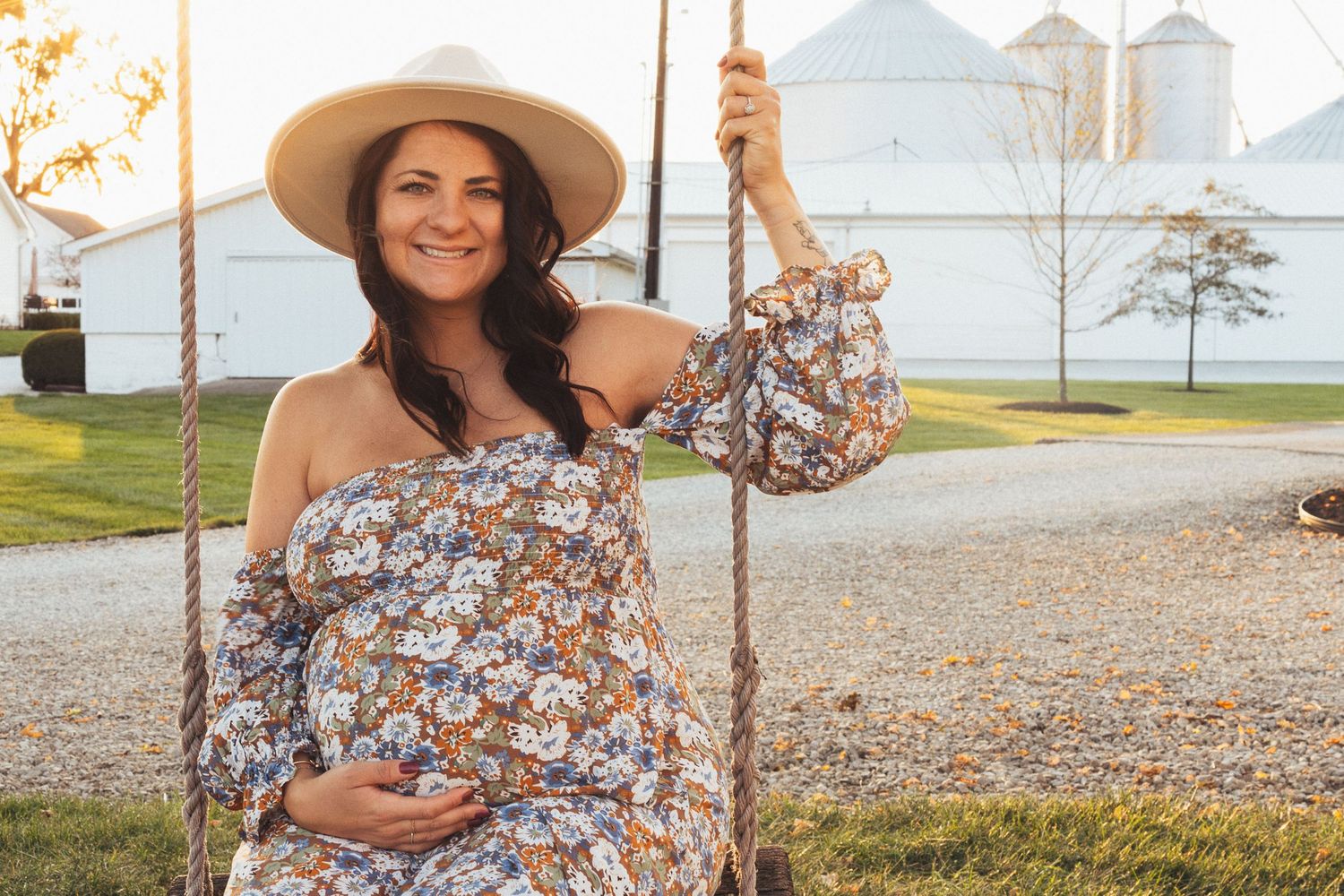 Woman in floral dress and wide brim hat poses in front of grain silos at golden hour.