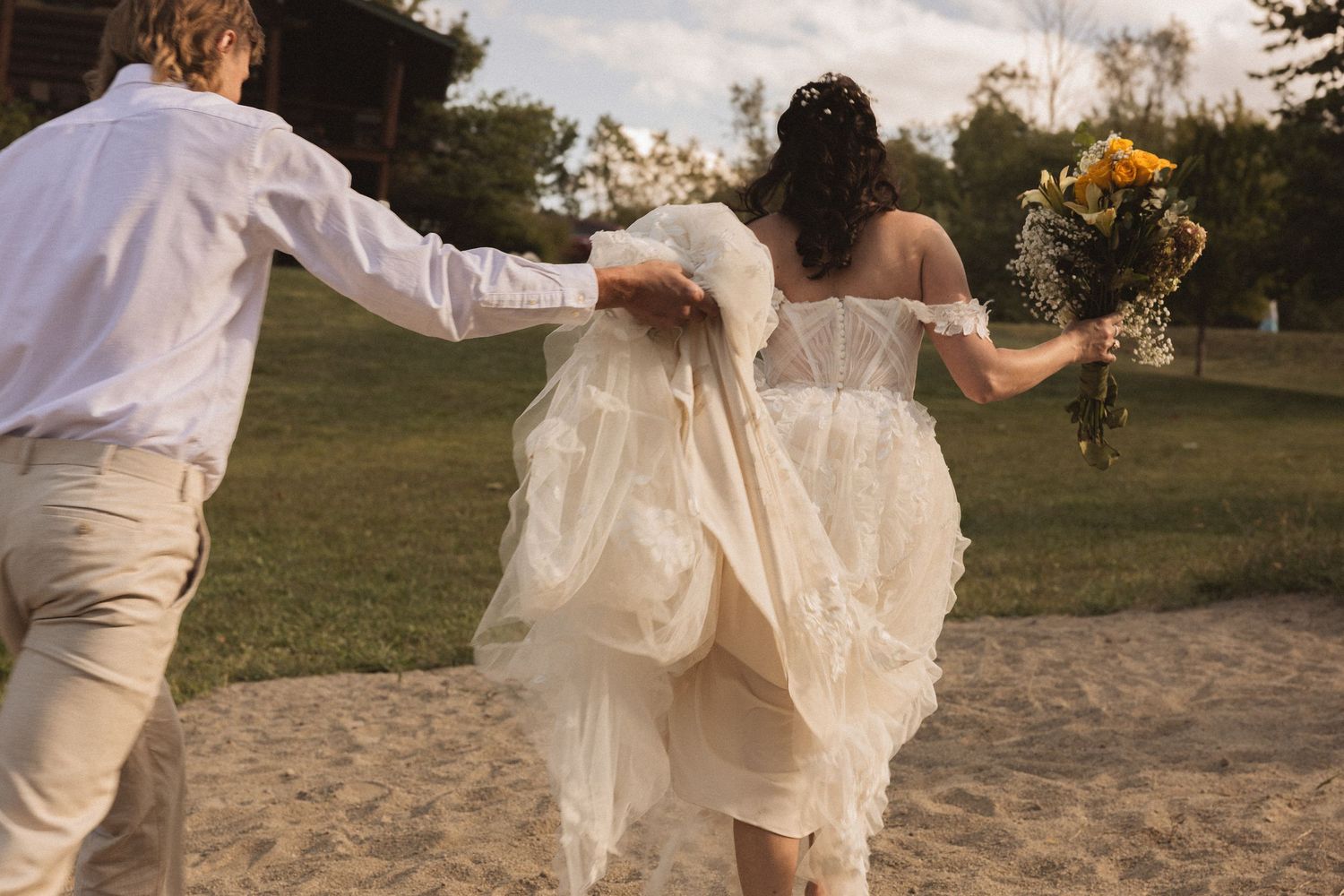 Two people holding hands and running through a field at sunset with flowing white dress and outfit in a romantic pose.
