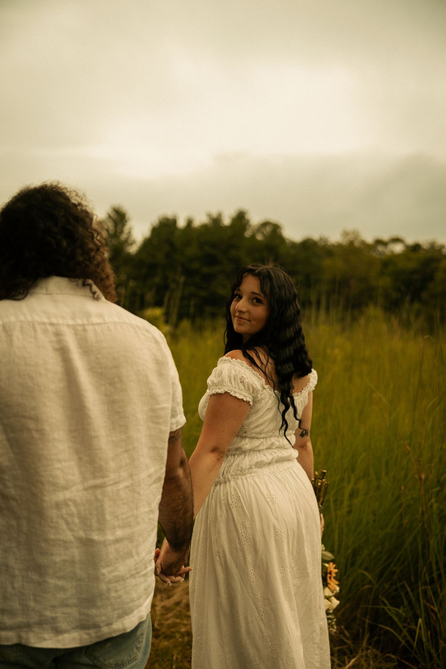 A woman in a white dress smiles over her shoulder at a man in a field with lush green trees under cloudy skies.