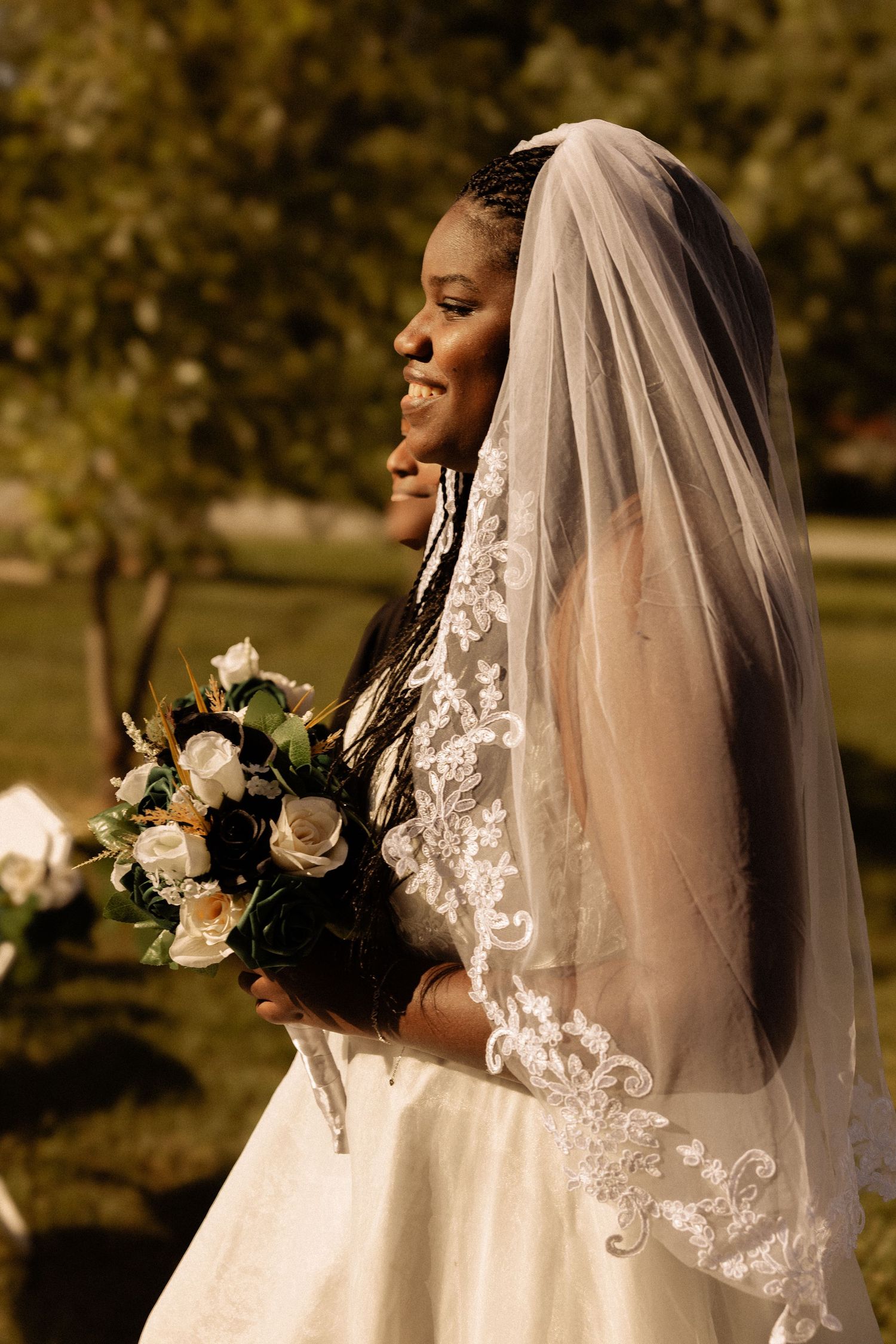 Beautiful bride in white gown and lace-trimmed veil smiles while holding cream and navy floral bouquet in garden setting.