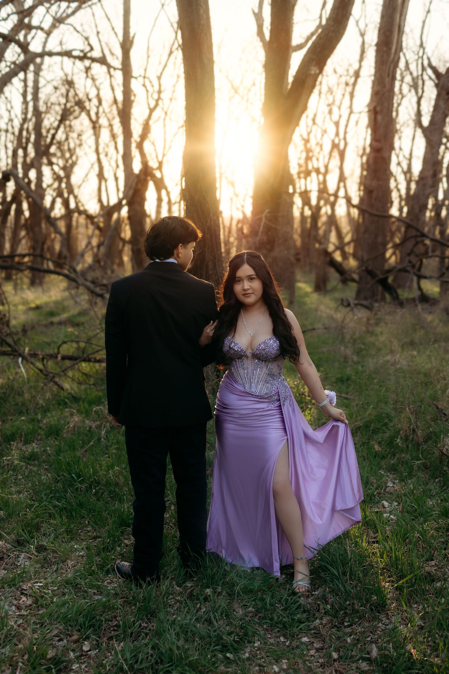 Couple in formal wear poses at sunset in a forest, woman in lavender satin gown with slit, man in black tuxedo.