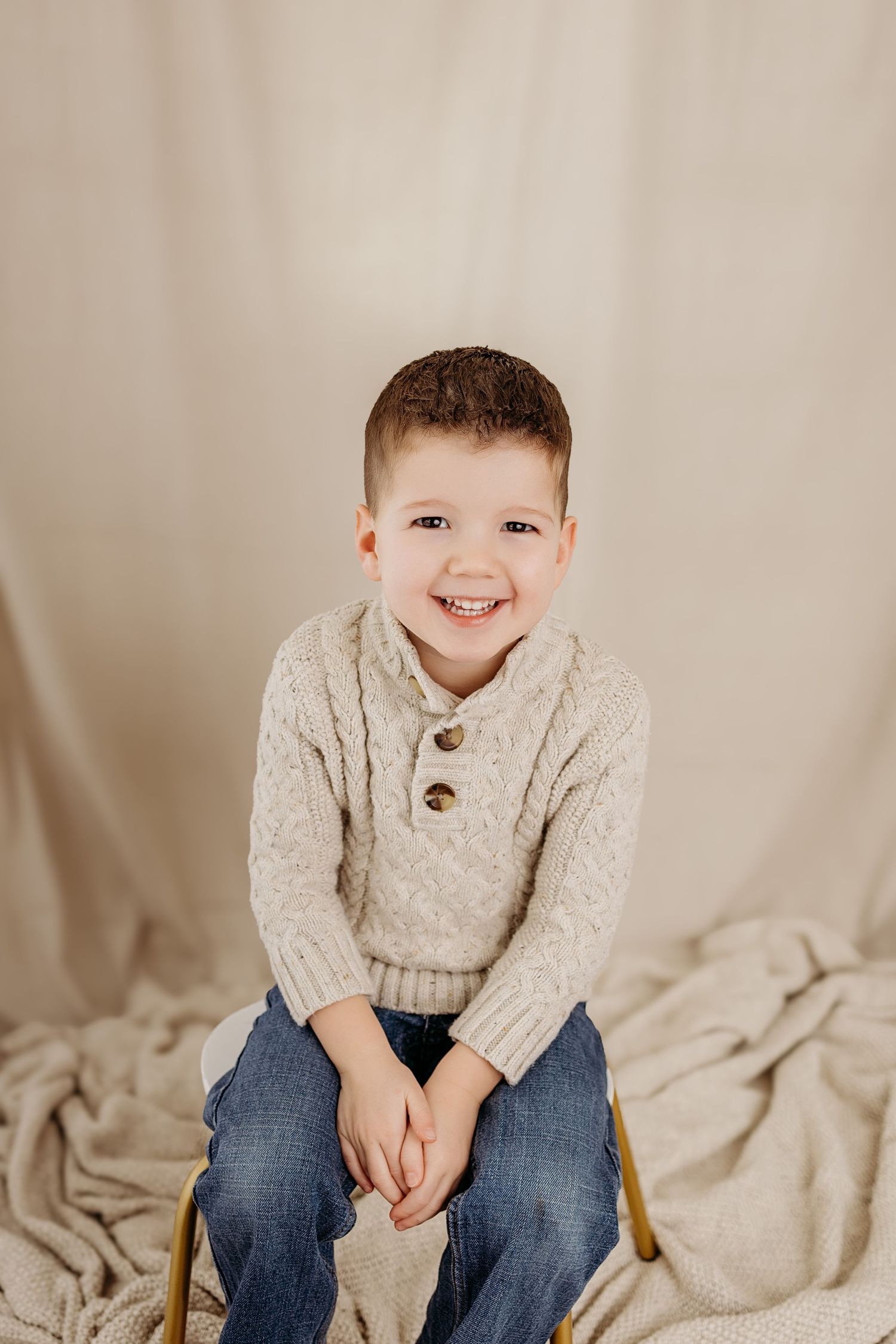 Cheerful young boy in cream knit sweater sitting and smiling against neutral backdrop.