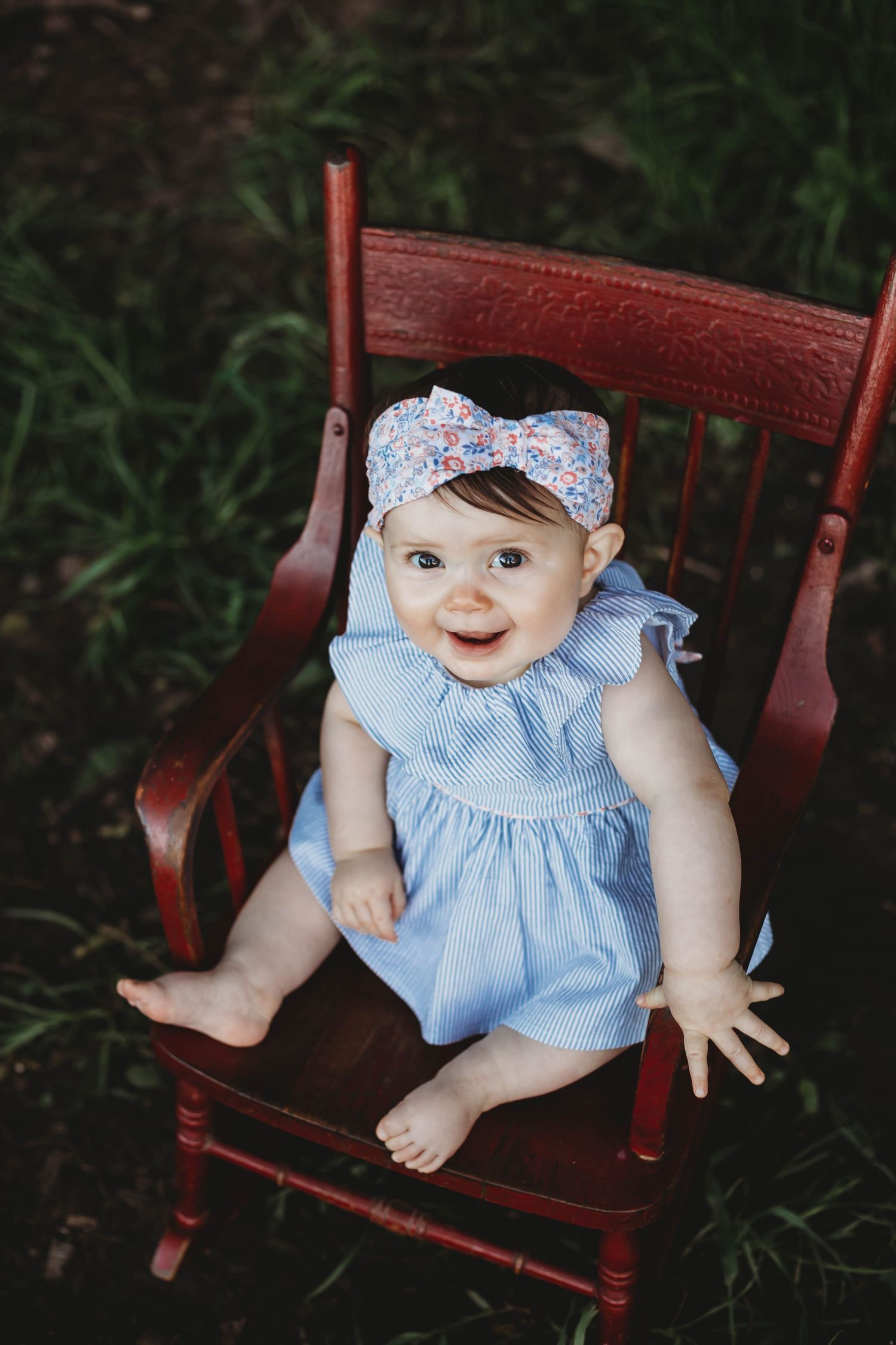 Smiling baby girl in blue dress and floral headband sitting in red wooden rocking chair outdoors.
