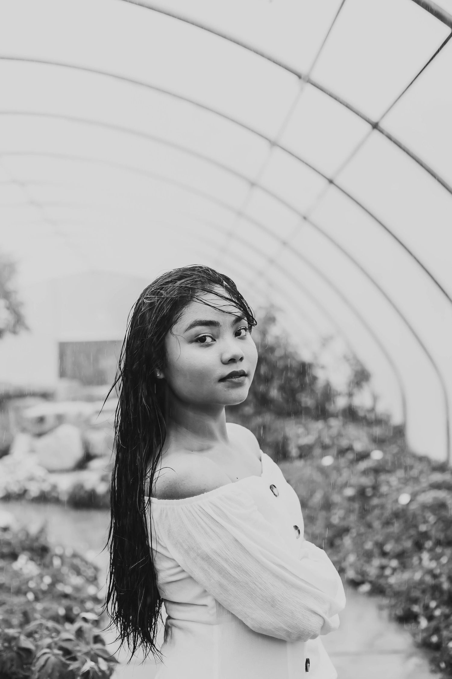 A young woman with long dark hair poses in a greenhouse in a black and white portrait photo.