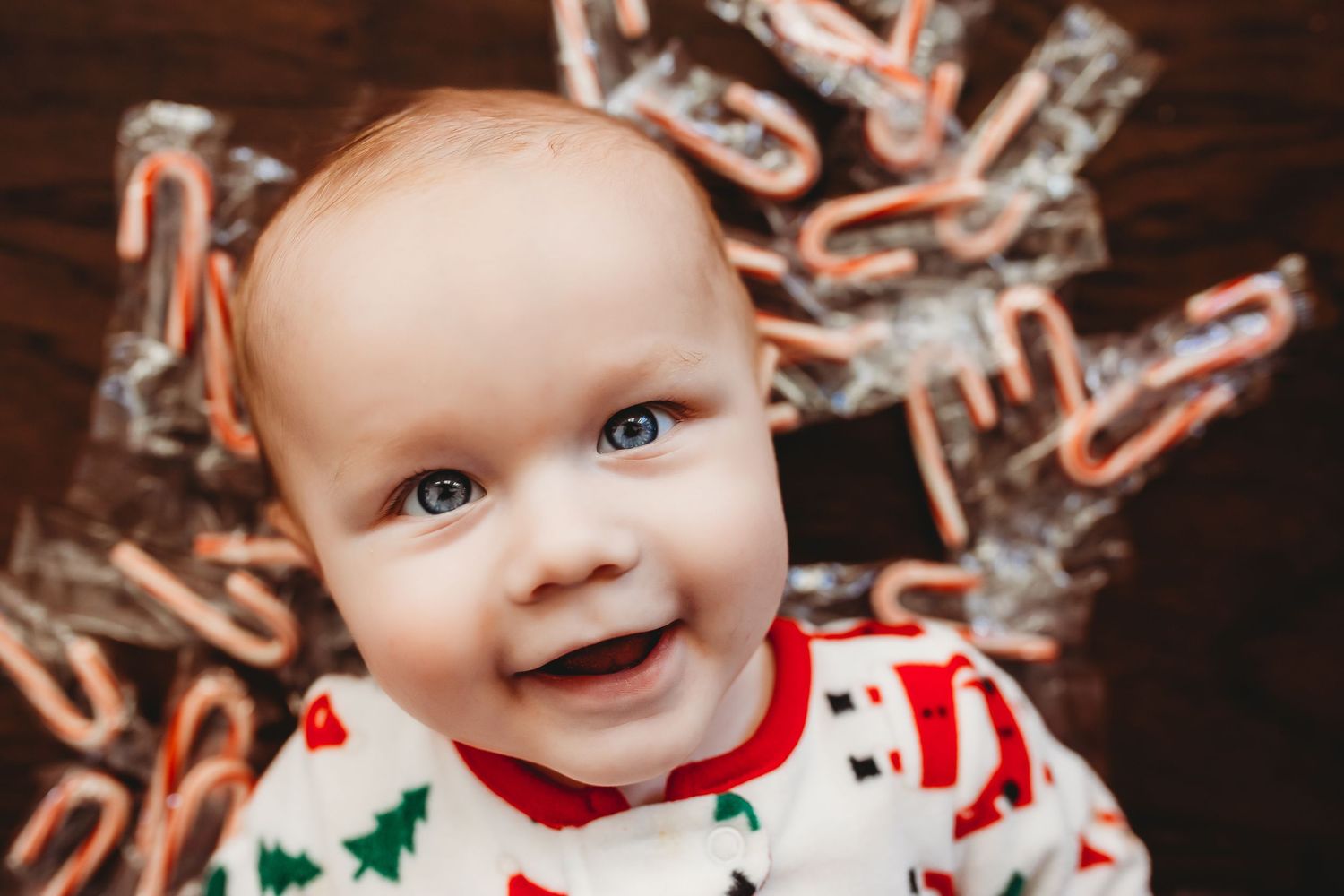 Happy baby in Christmas pajamas smiling surrounded by candy canes on a wooden surface.