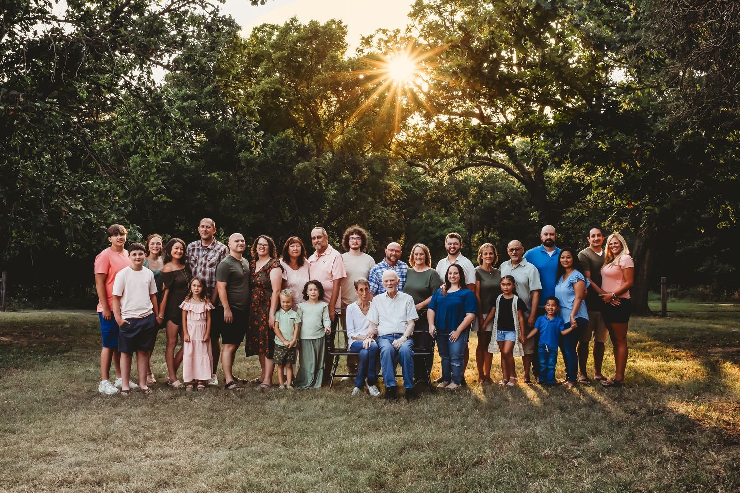 Large extended family group portrait outdoors at sunset surrounded by lush green trees.