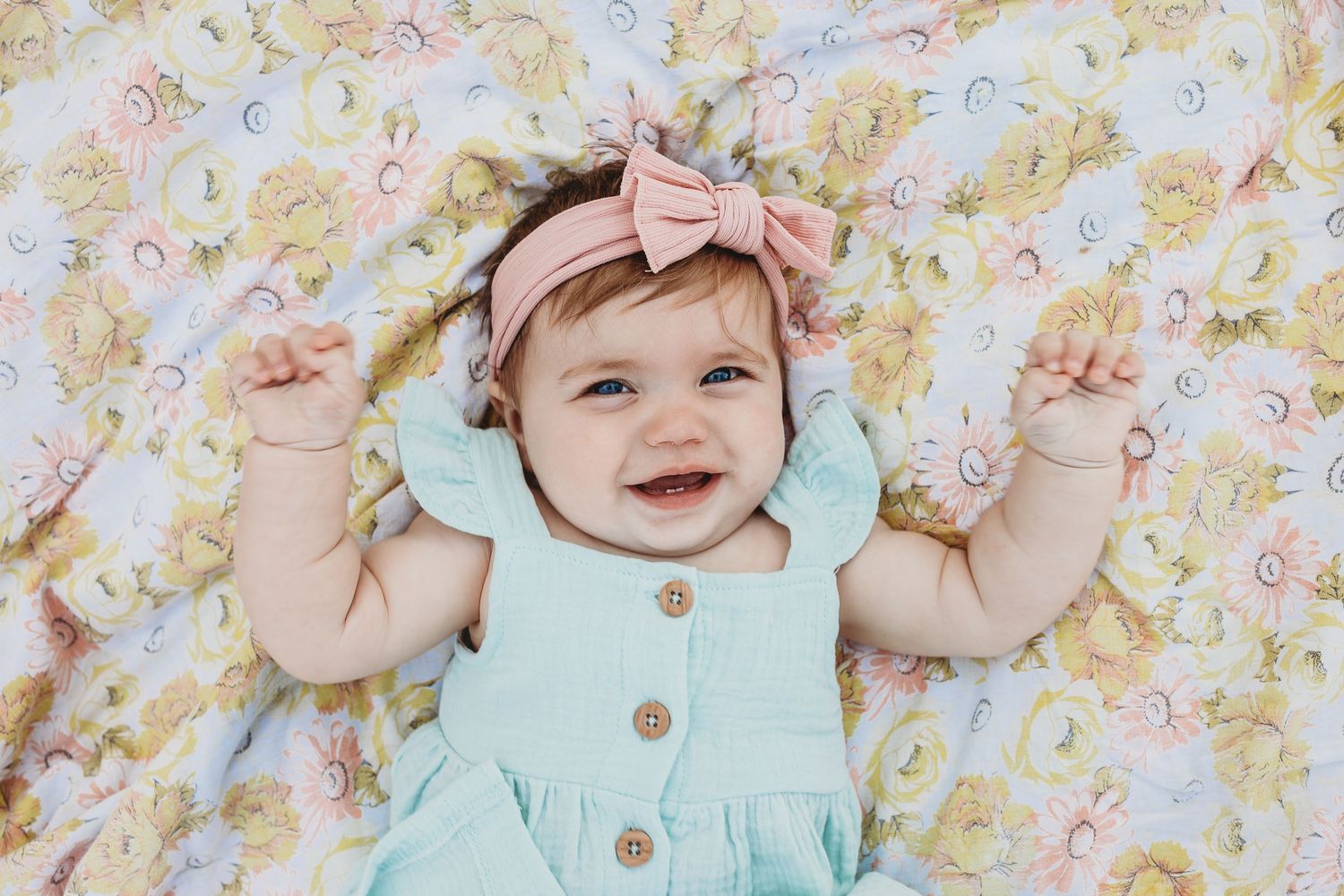 Happy baby girl in a mint dress smiling on a floral blanket wearing a pink bow headband.