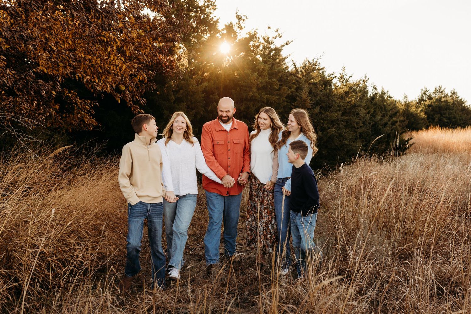 Family of six stands together smiling in tall golden grass field at sunset.