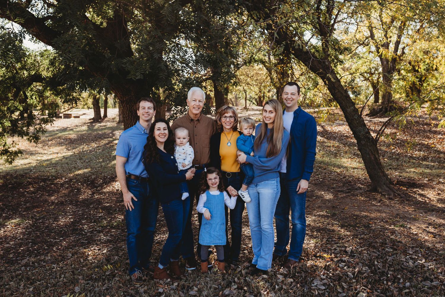 Extended family of nine posing together under large trees in autumn.