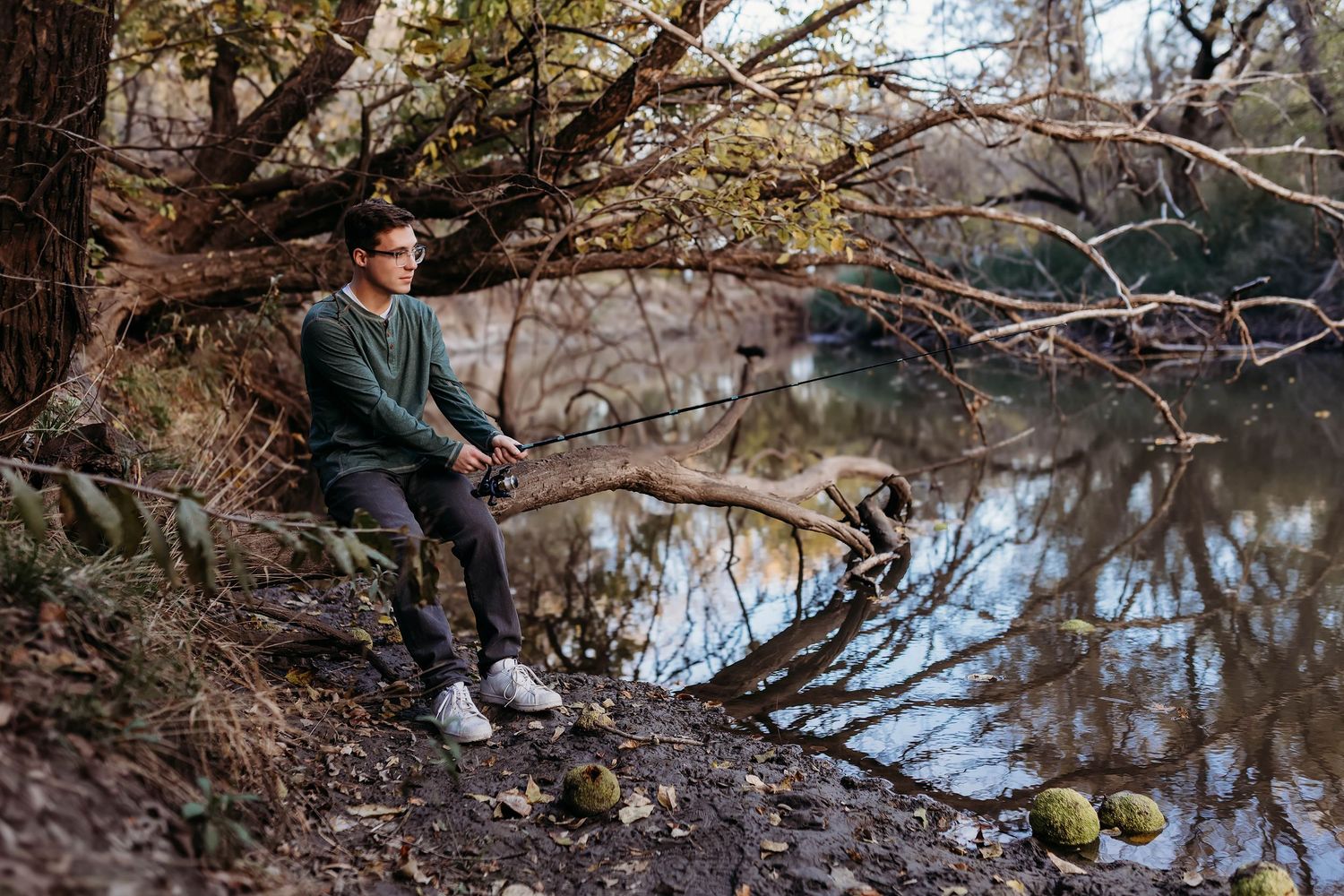 Young man sitting on a log by a calm river surrounded by autumn trees and foliage.