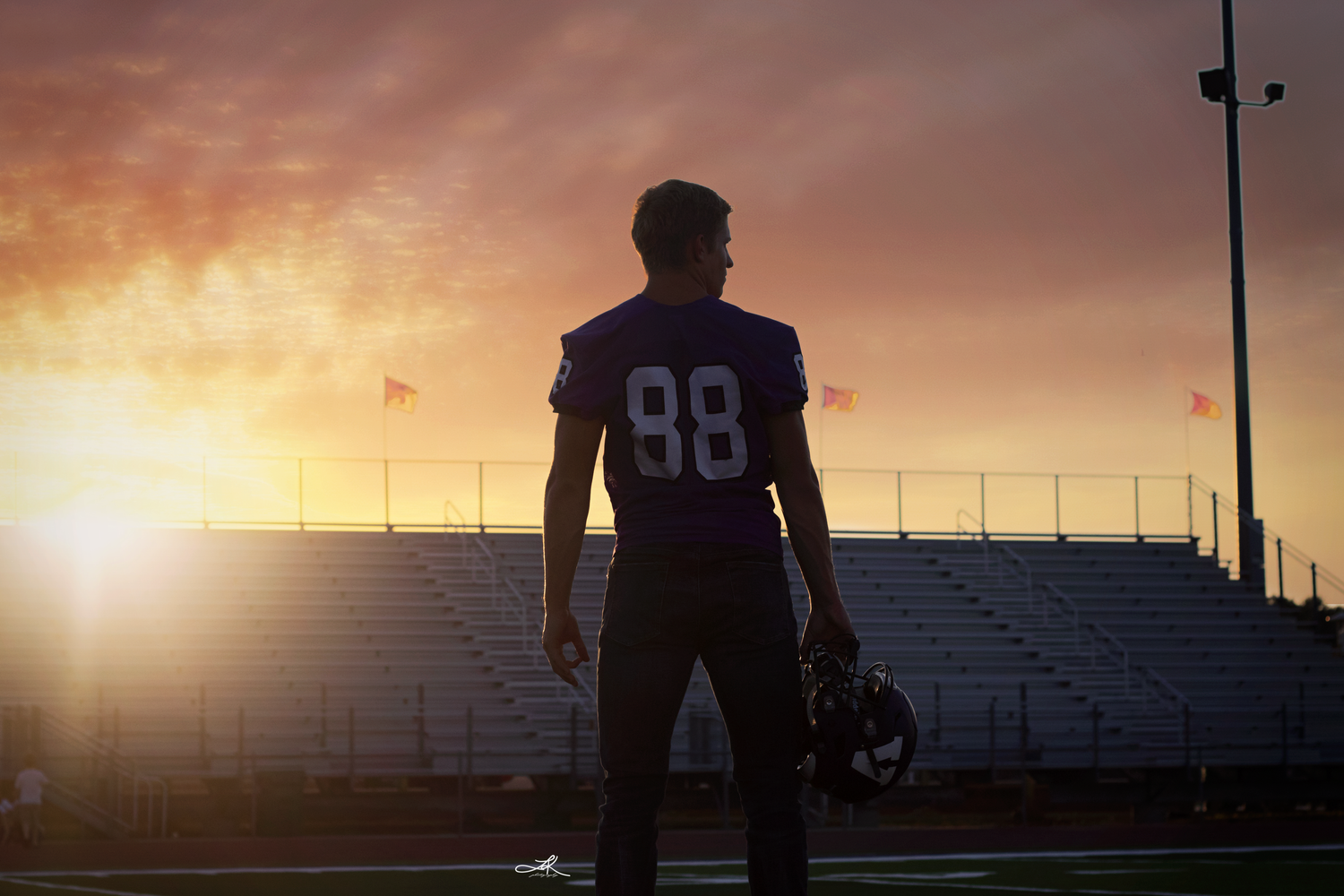 A football player wearing jersey number 88 stands silhouetted against a dramatic sunset sky on a stadium field.