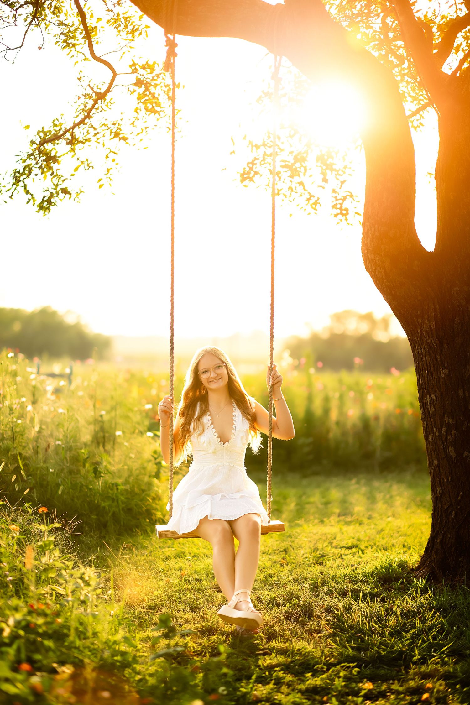 Young girl in white dress sitting on a rope swing under a tree, bathed in golden sunset light in a green meadow.