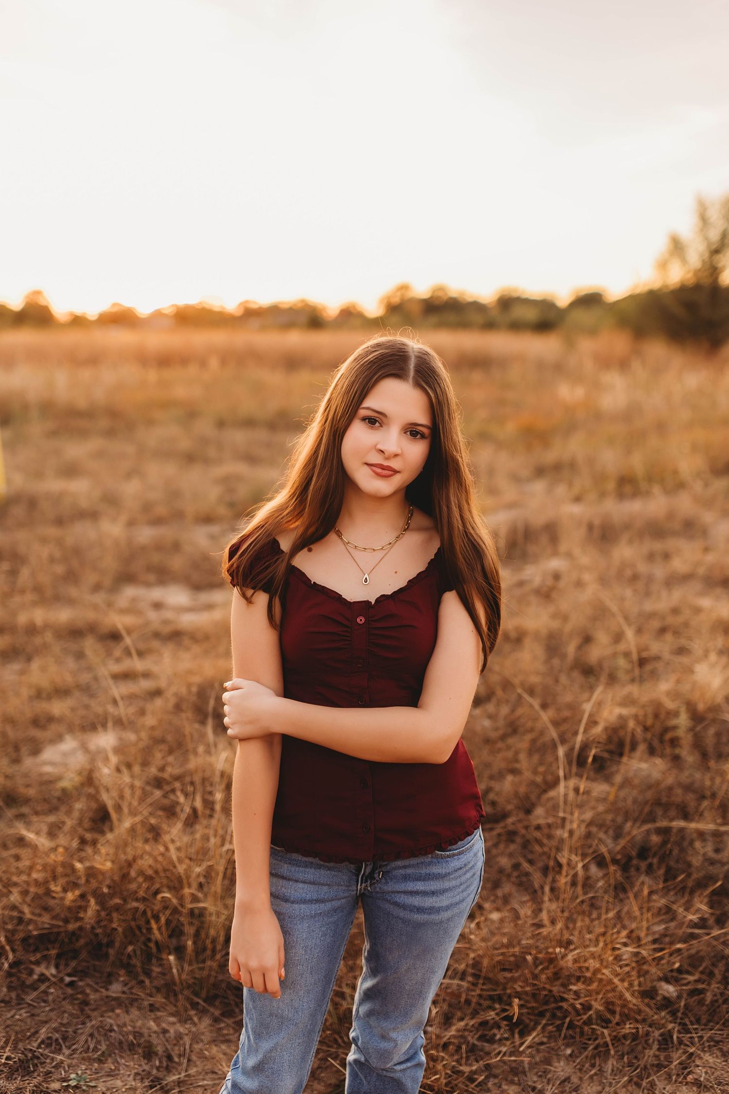 Young woman in burgundy top and jeans poses in golden field during sunset, long brown hair, wearing layered necklaces.