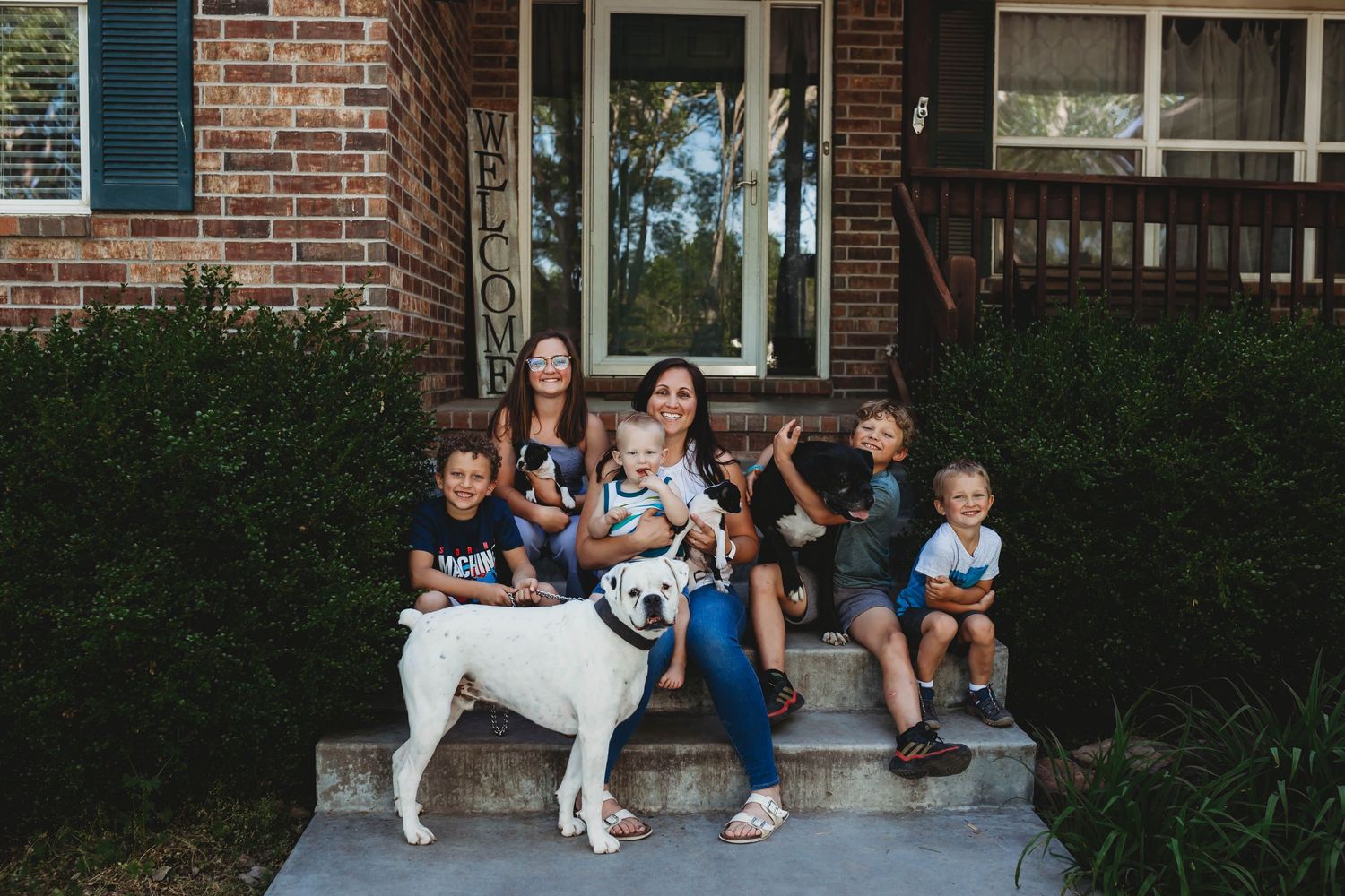 Family with four kids and white bulldog sitting on brick home front steps outdoors.