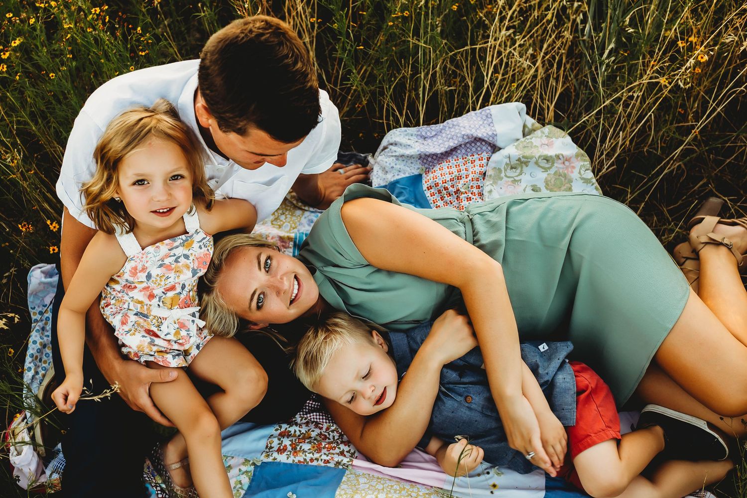 Playful family of four laughing and cuddling together on a quilt in a meadow.