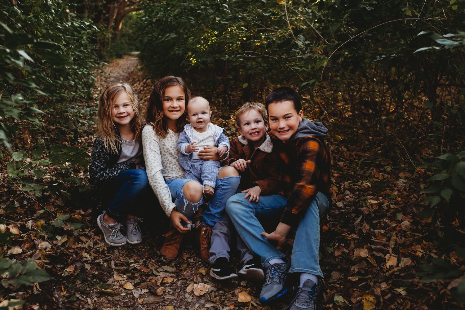 Five children smiling together among autumn leaves in a lush green forest setting.