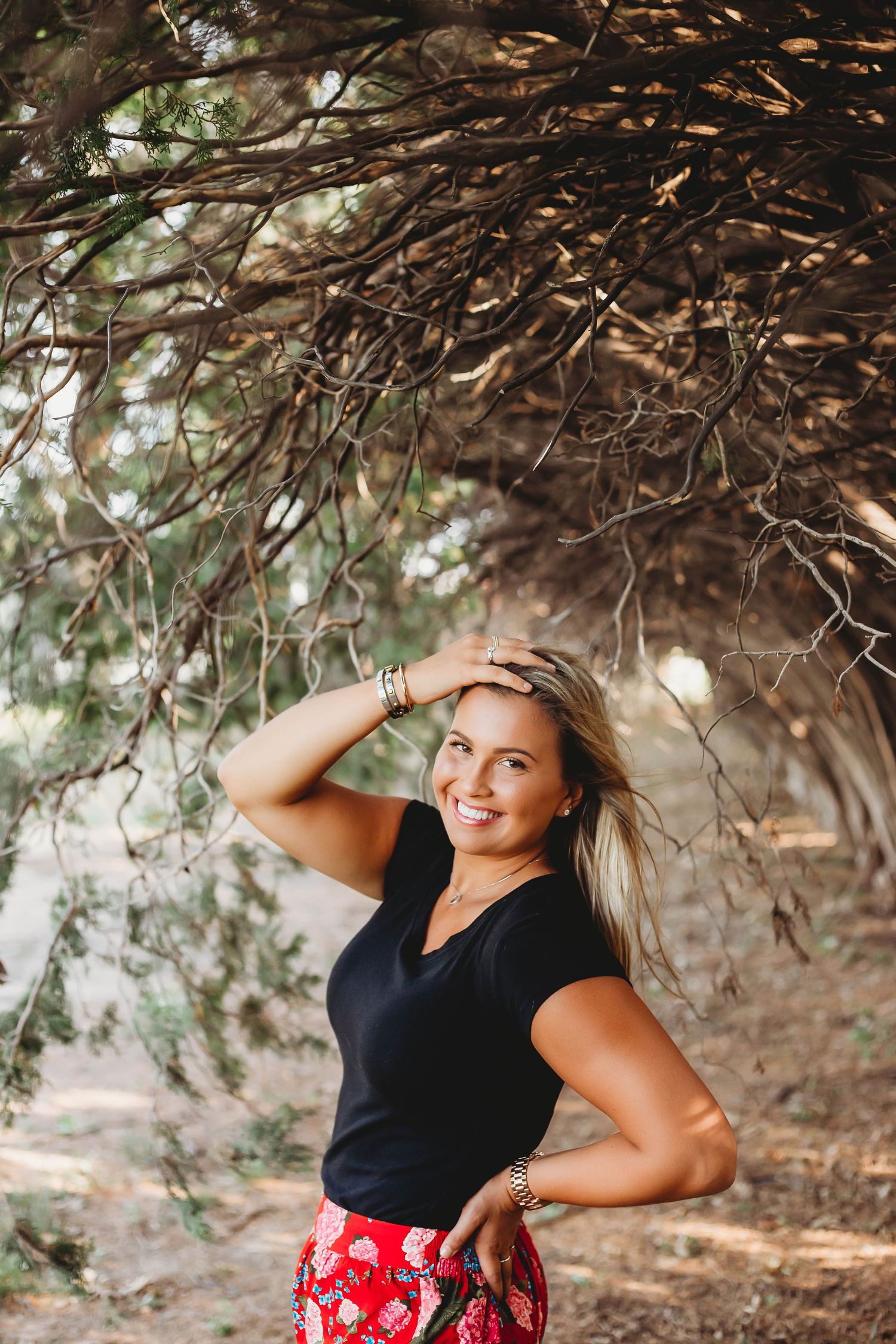Smiling blonde woman in black top and red floral skirt posing under arching tree branches outdoors.