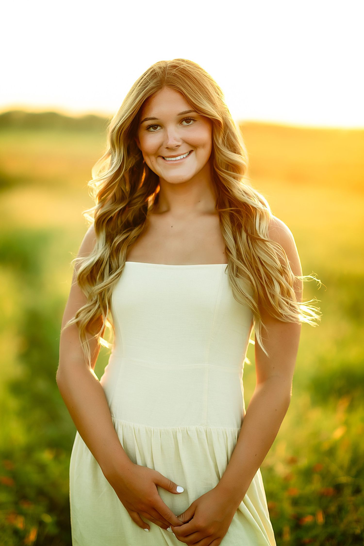 Young blonde woman in white sundress smiling outdoors in golden sunset light in a green field.