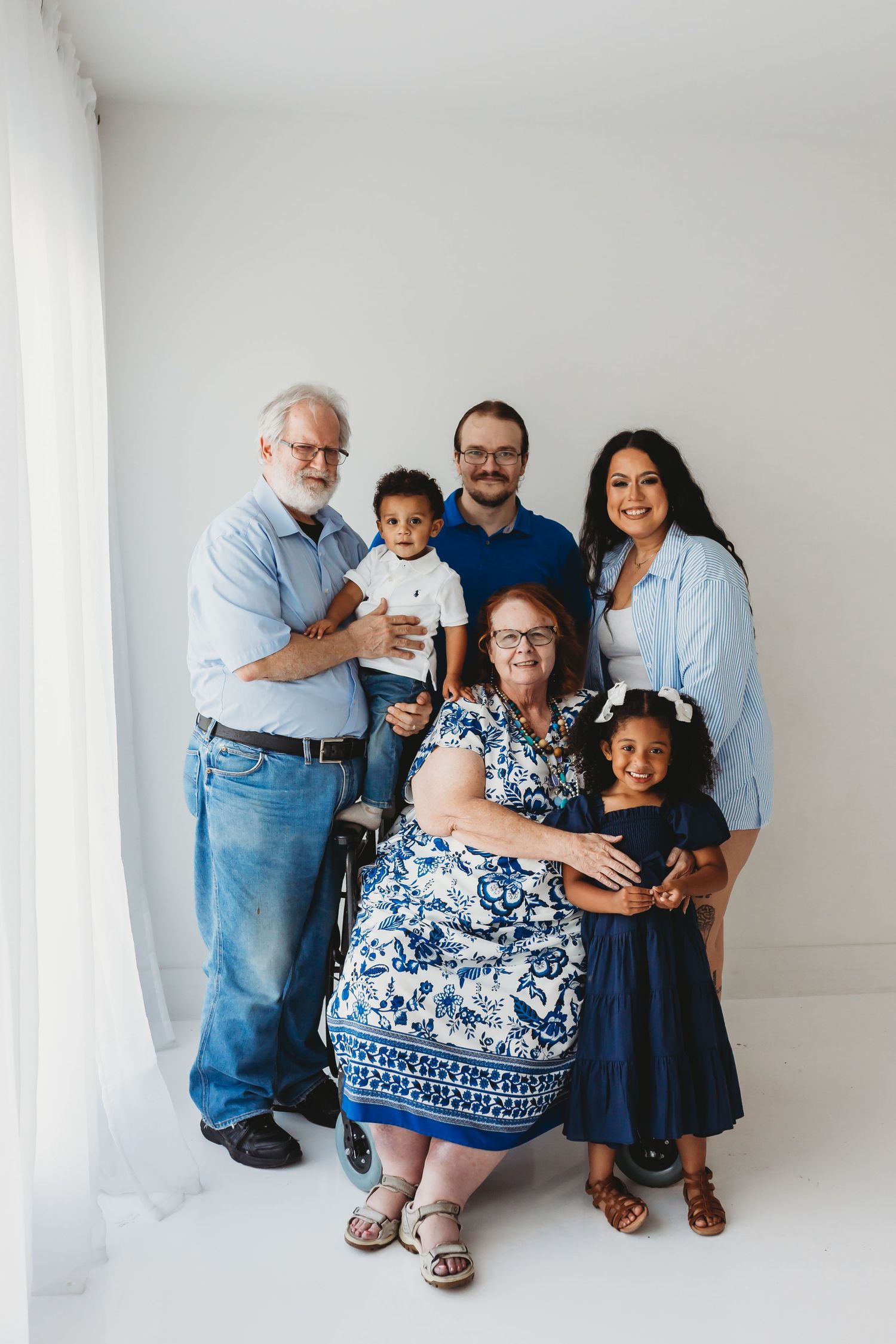 Multi-generational family portrait with grandparents, parents, and two young children posing together in a bright white studio.