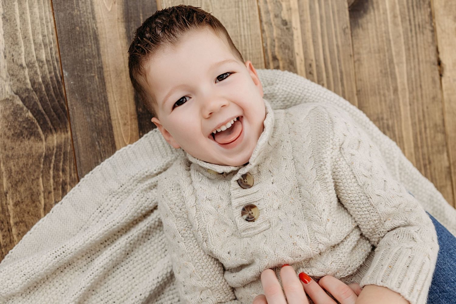 Laughing toddler in cream knit sweater being held against a rustic wood background.