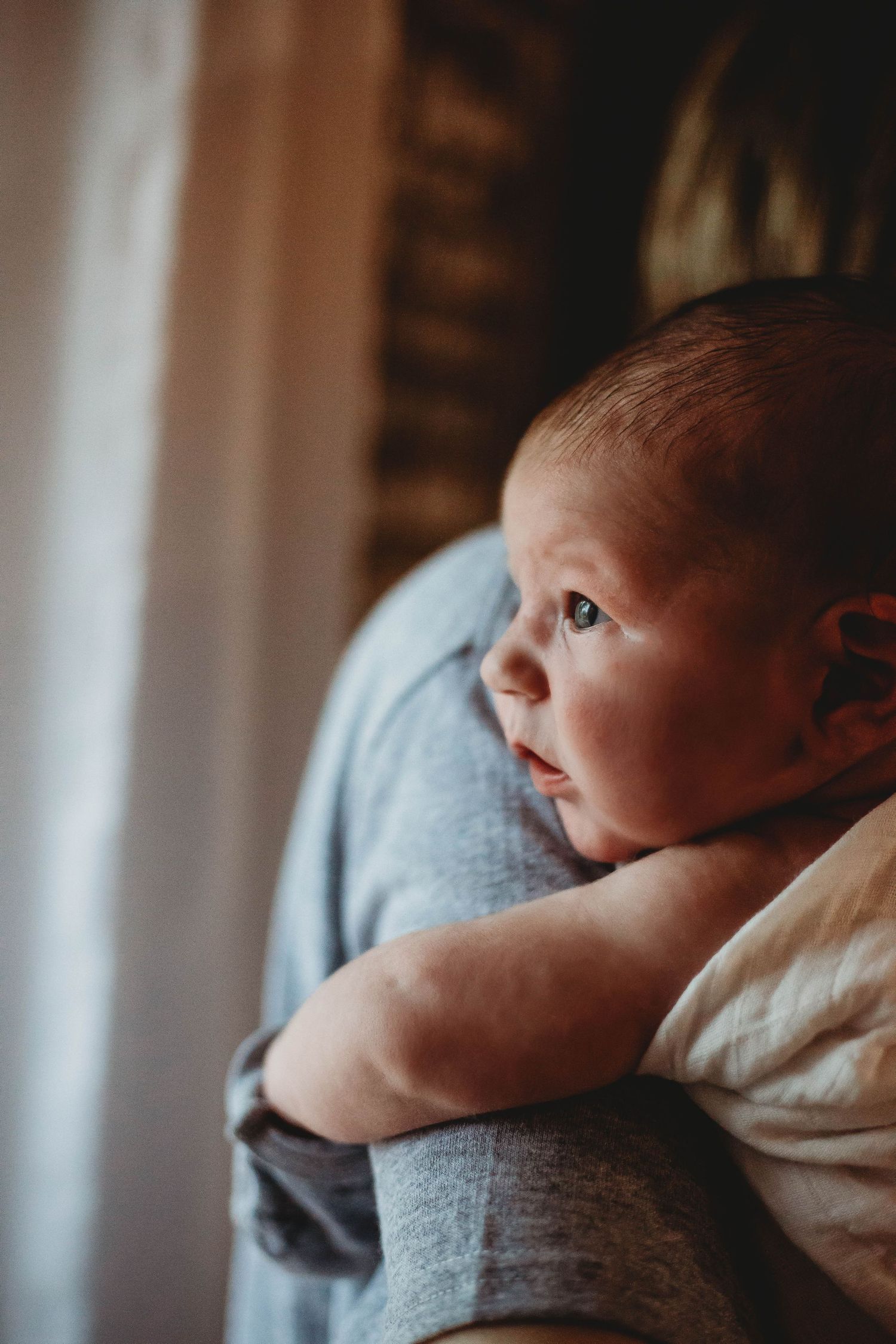 A newborn baby resting on an adult's shoulder, gazing curiously to the side in a softly lit room.