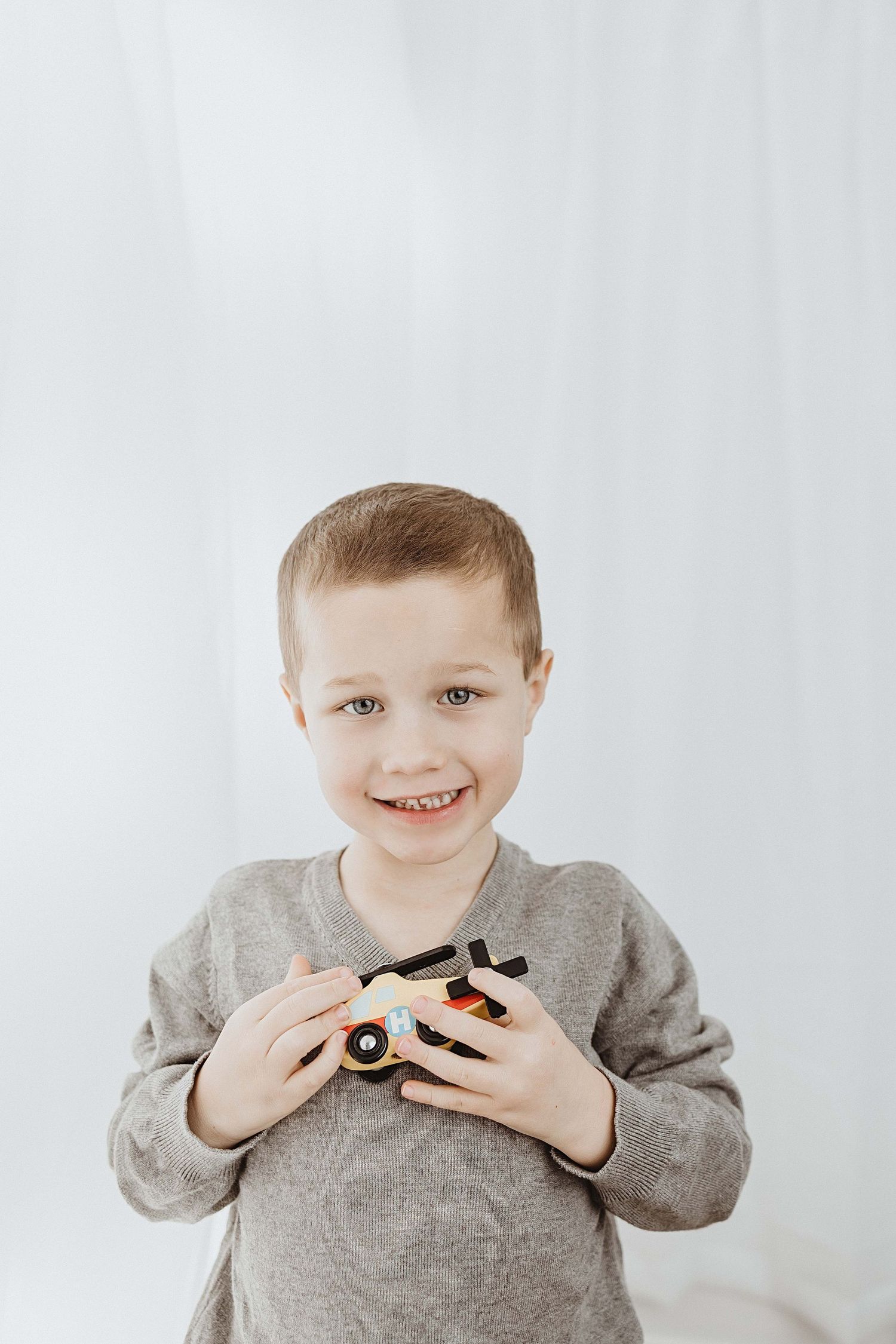 Smiling young boy in gray sweater holding a wooden toy camera against a white background.