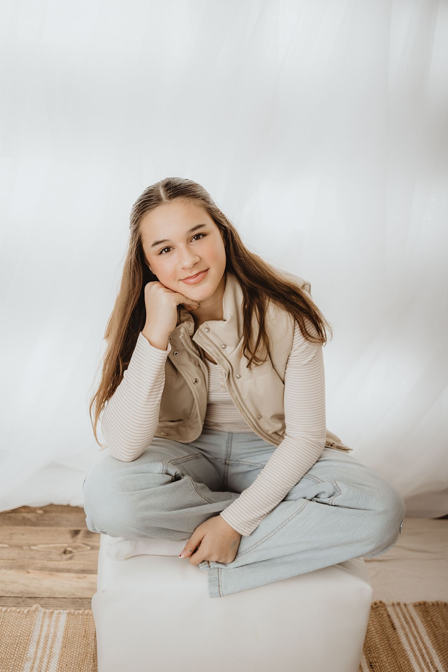 Teenage girl in beige vest and jeans sitting cross-legged on white ottoman against white backdrop.