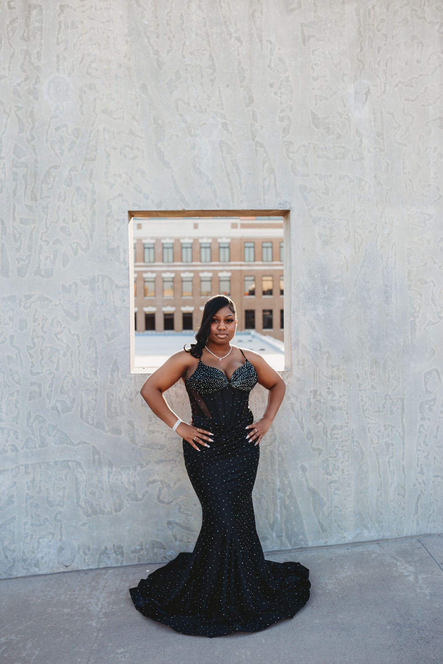 A woman in an elegant black sequined mermaid gown poses confidently against a light stone wall outdoors.