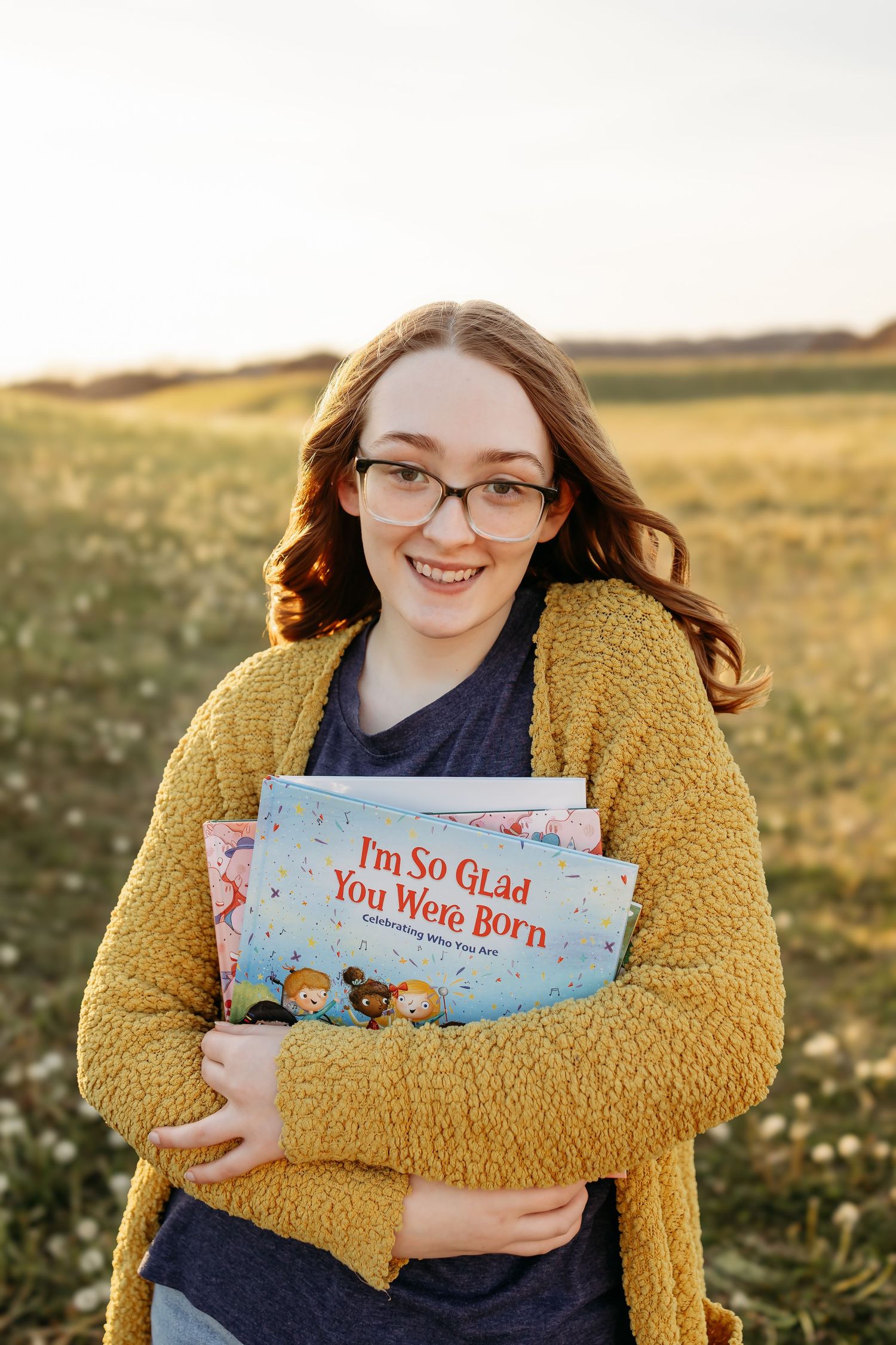 Smiling young woman in yellow cardigan holding children's book I'm So Glad You Were Born outdoors.
