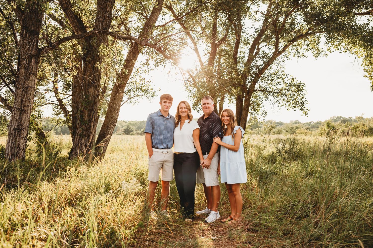 A family of four poses together under sunlit trees in a golden meadow during an outdoor portrait session.