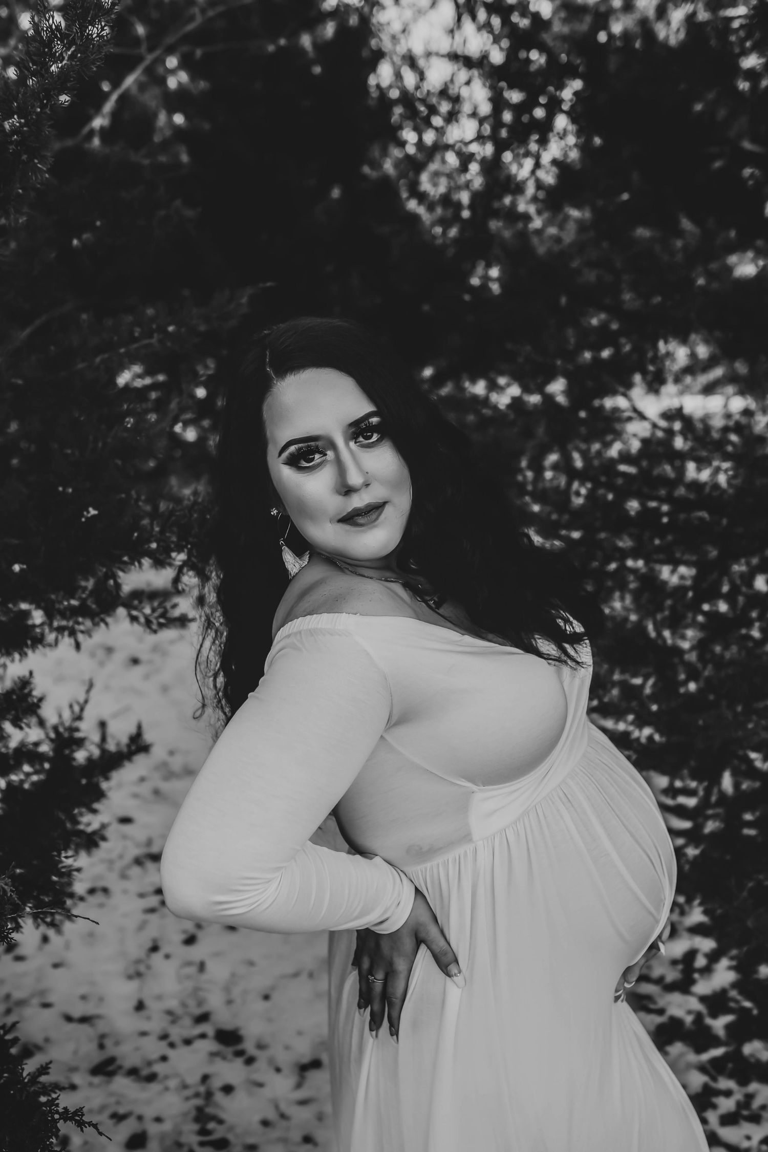 Black and white portrait of a pregnant woman in a white gown posing gracefully among snow-dusted evergreen trees.