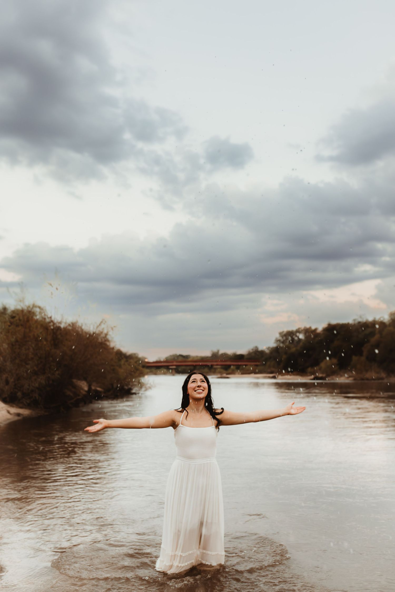 Joyful young woman in white dress standing in a river with arms outstretched under cloudy sky.