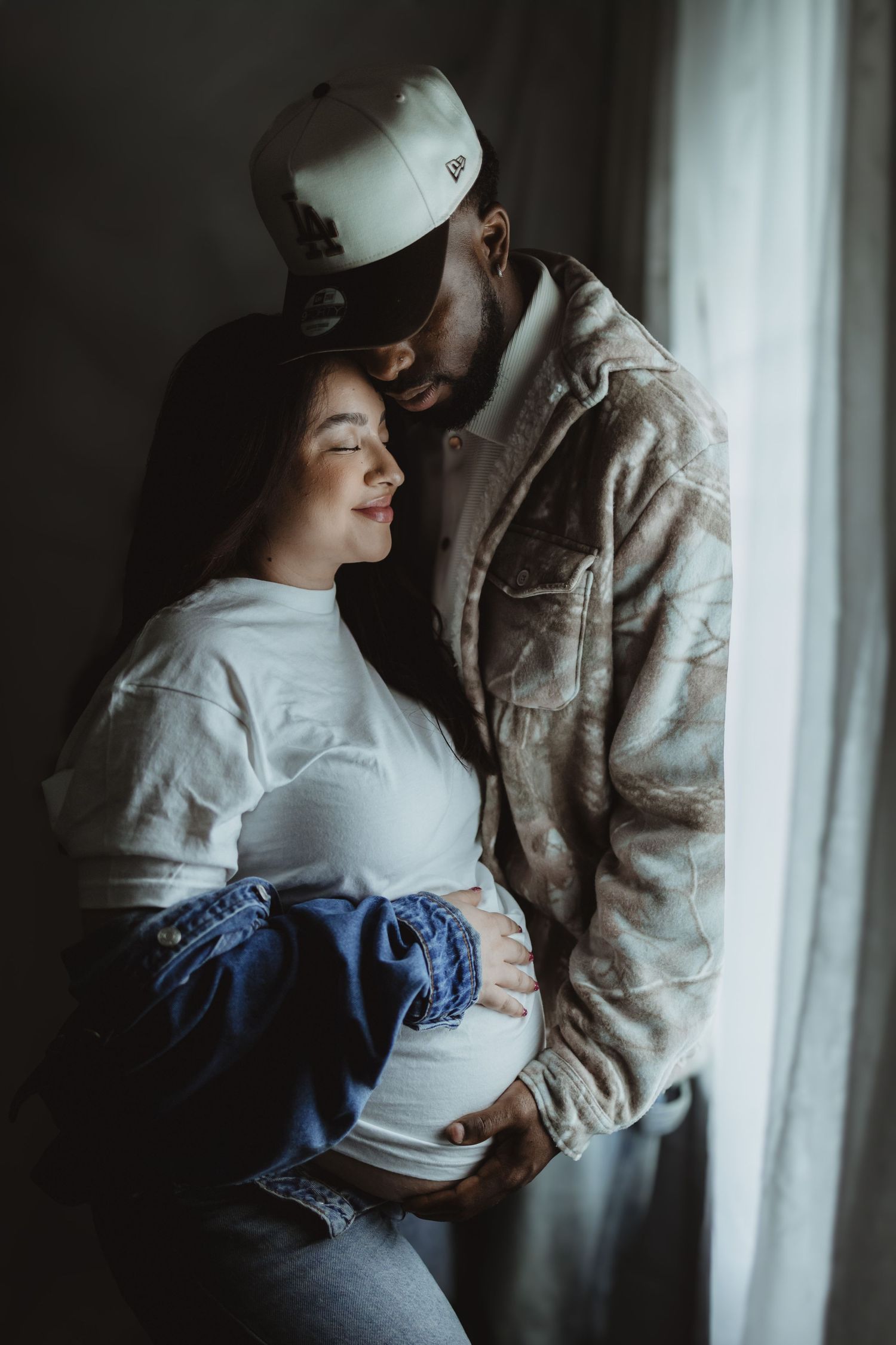 Intimate maternity portrait of interracial couple embracing by window, man tenderly holds pregnant womans belly.