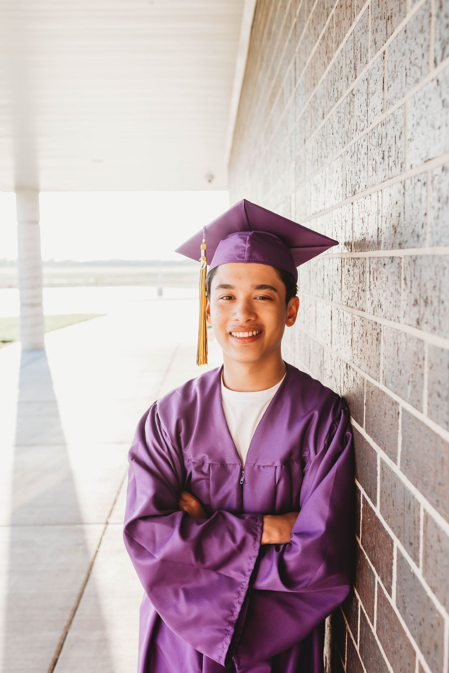 Smiling graduate in purple cap and gown leaning against brick wall with arms crossed.