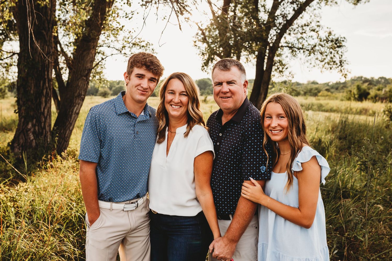 Happy family of four posing together in golden sunlit field with trees in background.