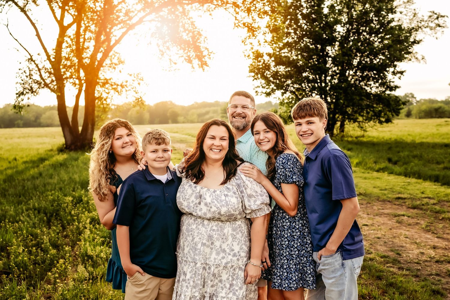 Family of six smiling together outdoors at golden hour in a green field with trees and warm sunlight.