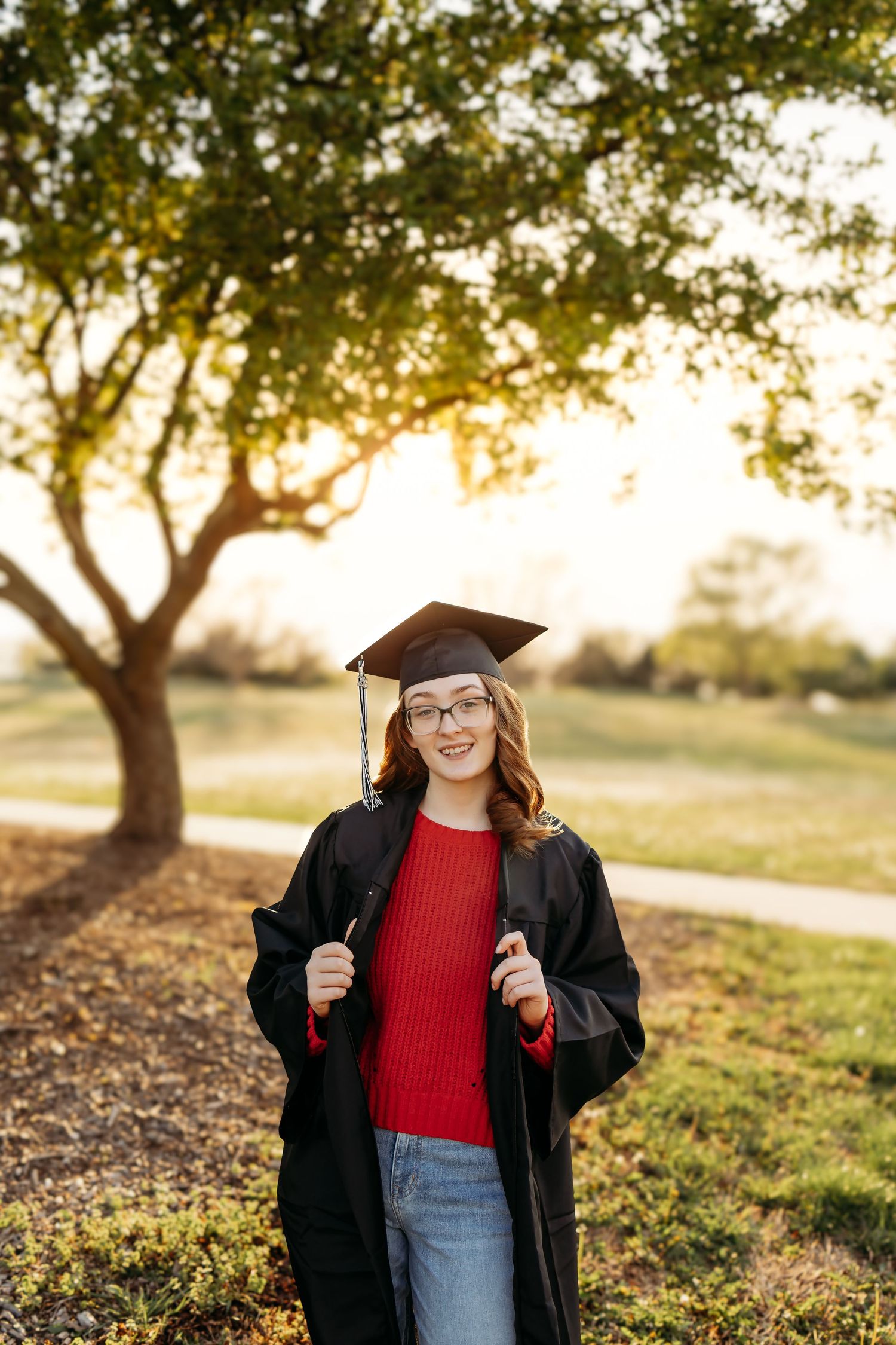 Young woman in black graduation cap and gown smiling outdoors under golden sunlight trees.