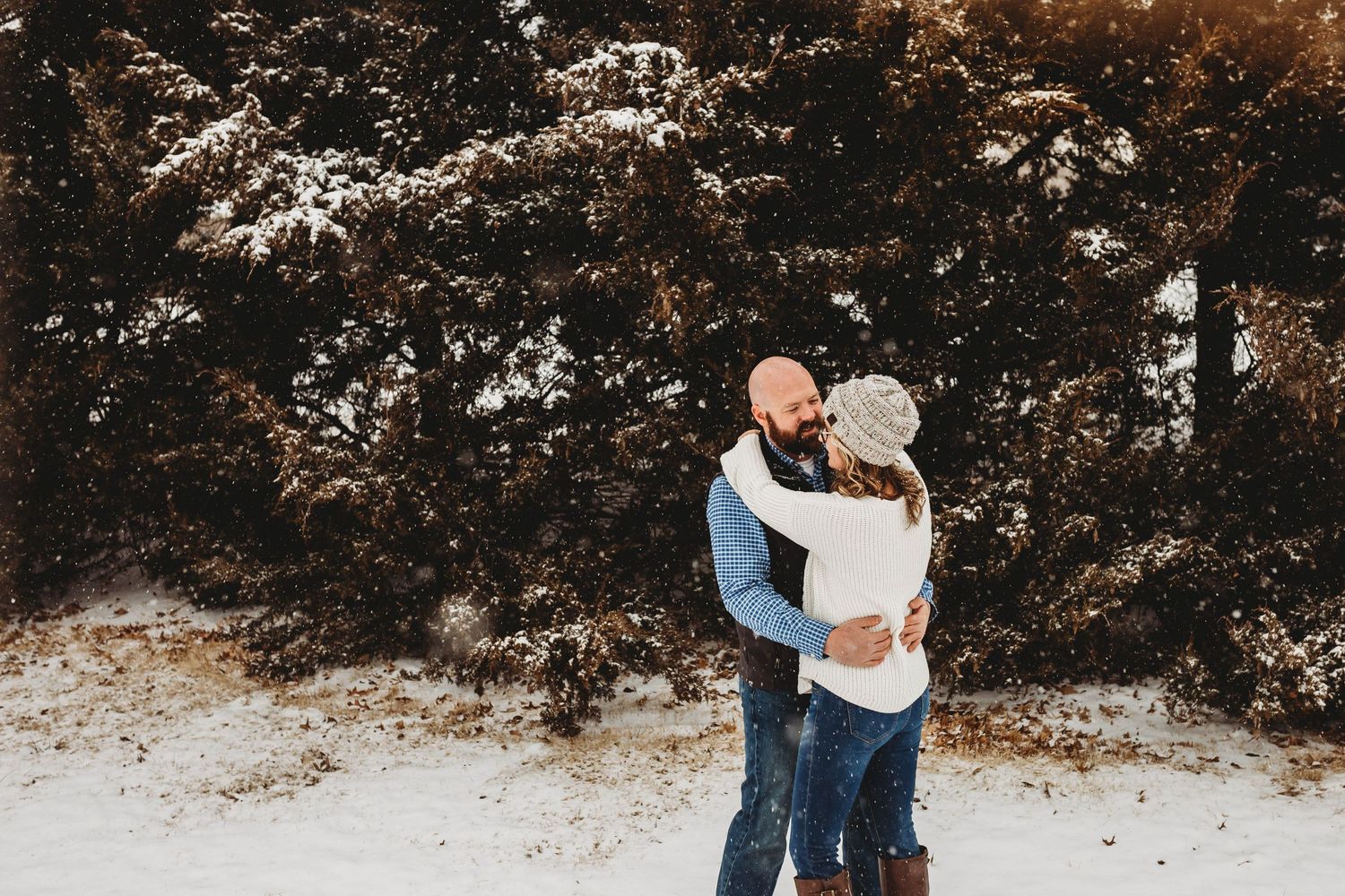 Couple embracing in a snowy winter landscape with snow-covered evergreen trees in the background.