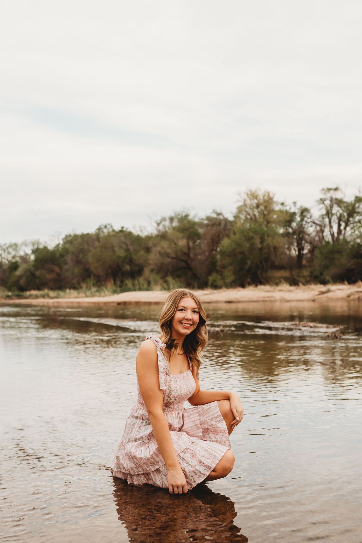A smiling young woman in a pink floral dress crouches in a shallow river with trees in the background.