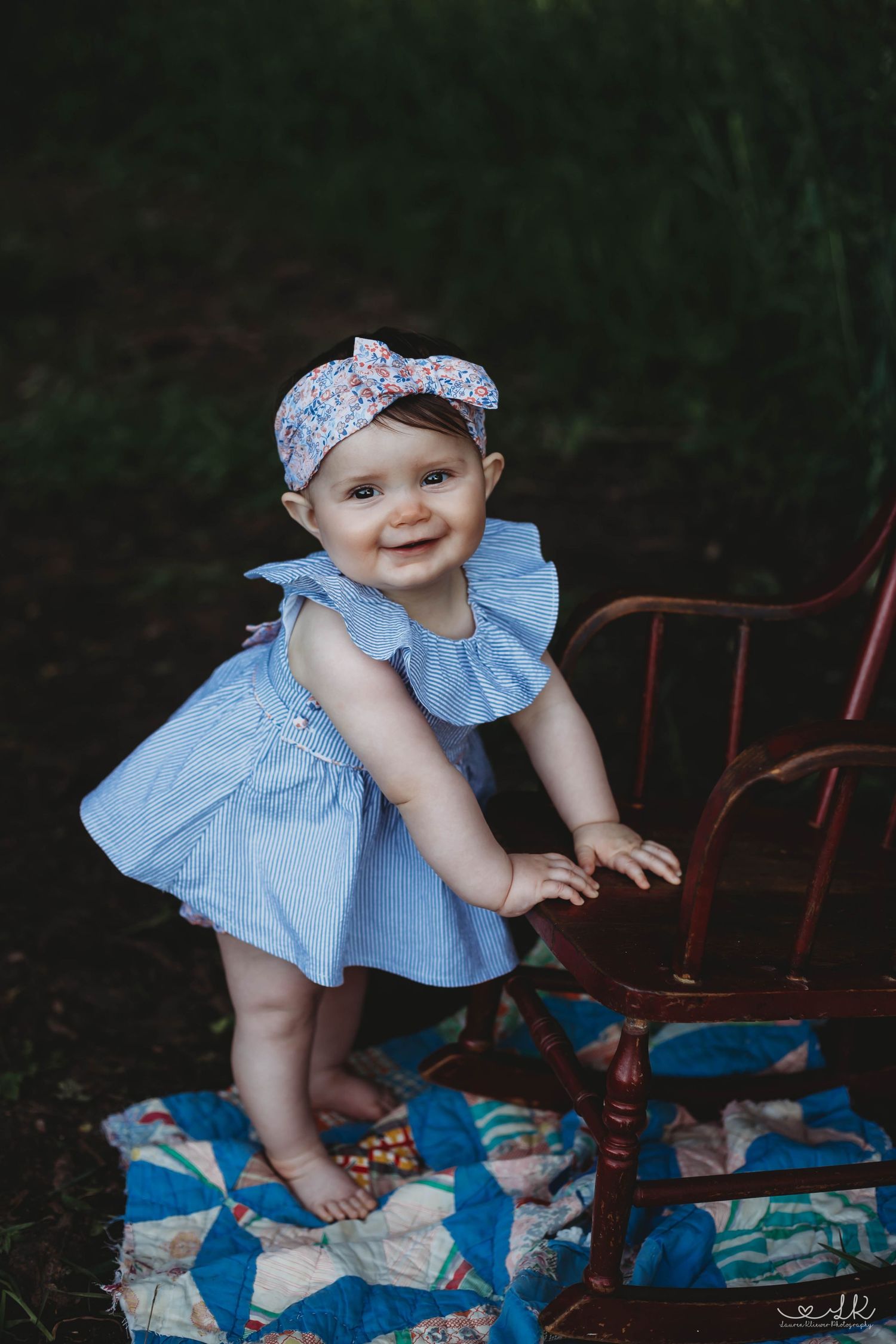 A happy baby girl in a blue ruffle dress and floral headband stands beside a red chair on a quilt outdoors.