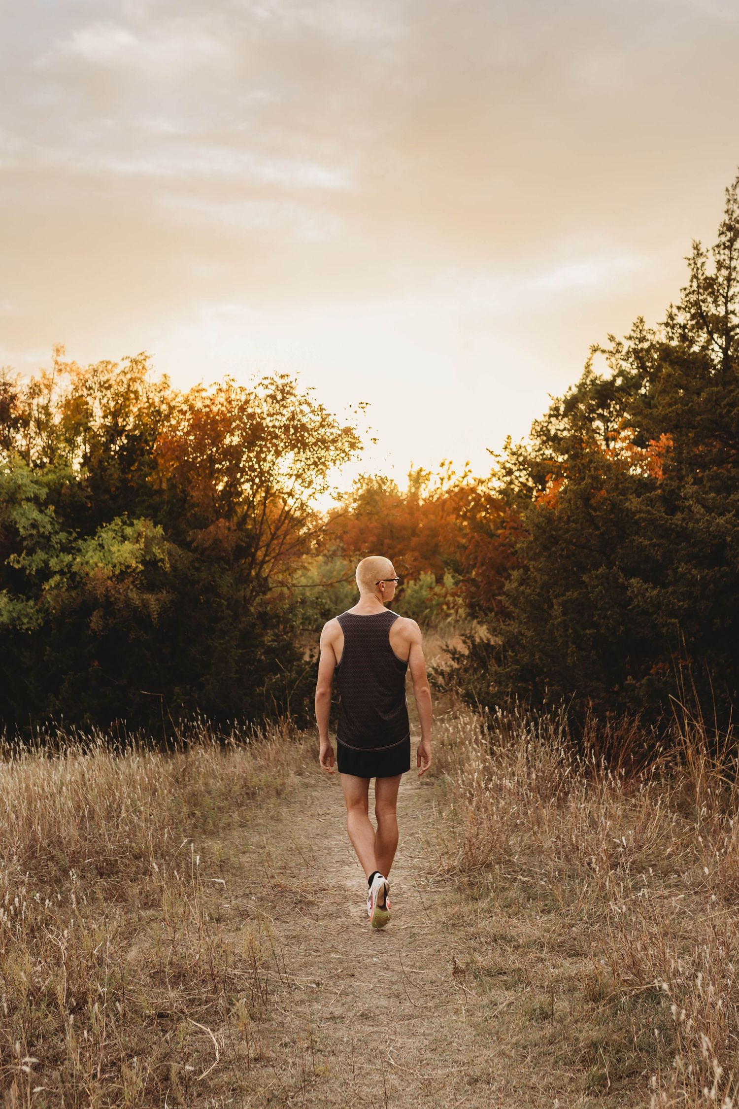 A man in a black tank top and shorts walks along a dirt trail surrounded by golden grass and trees at sunset.