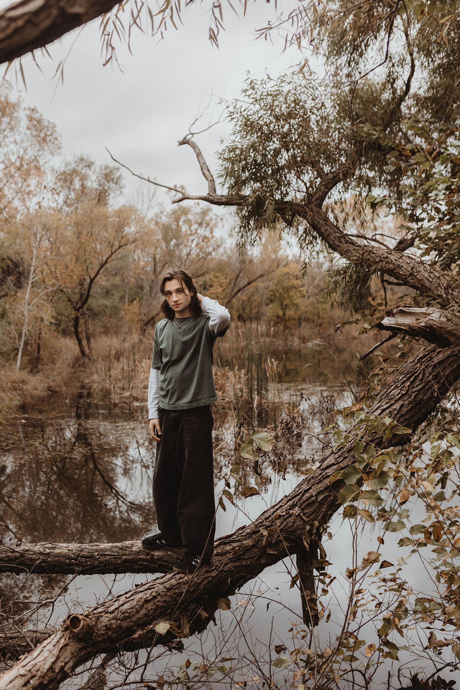Young person in green sweatshirt standing on a fallen tree by a reflective autumn pond.