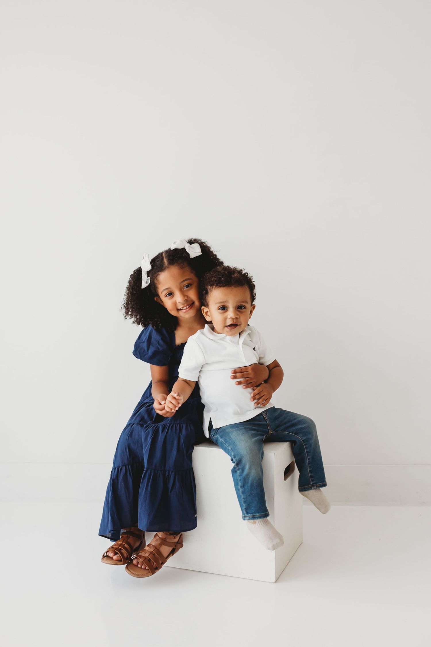 Young girl in navy dress hugging her little brother on a white cube.