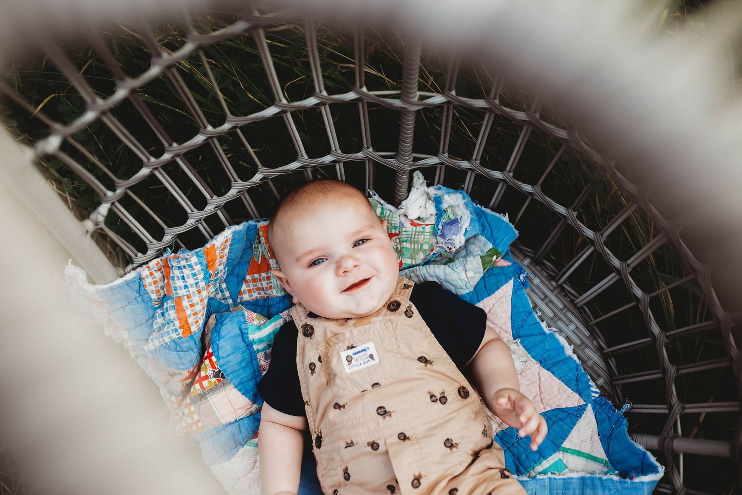 Smiling baby in tan overalls lying in a wicker basket with colorful quilt, viewed from above.