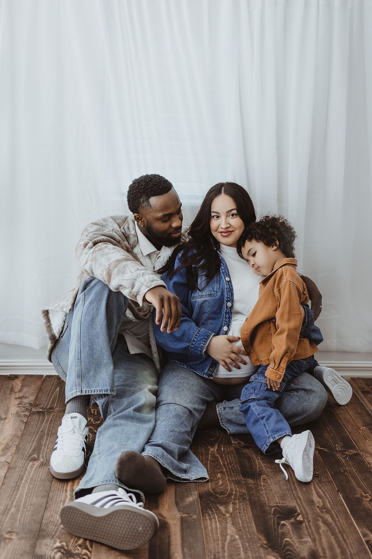 A multiracial family with a young child sits together on a wooden floor against white curtains.