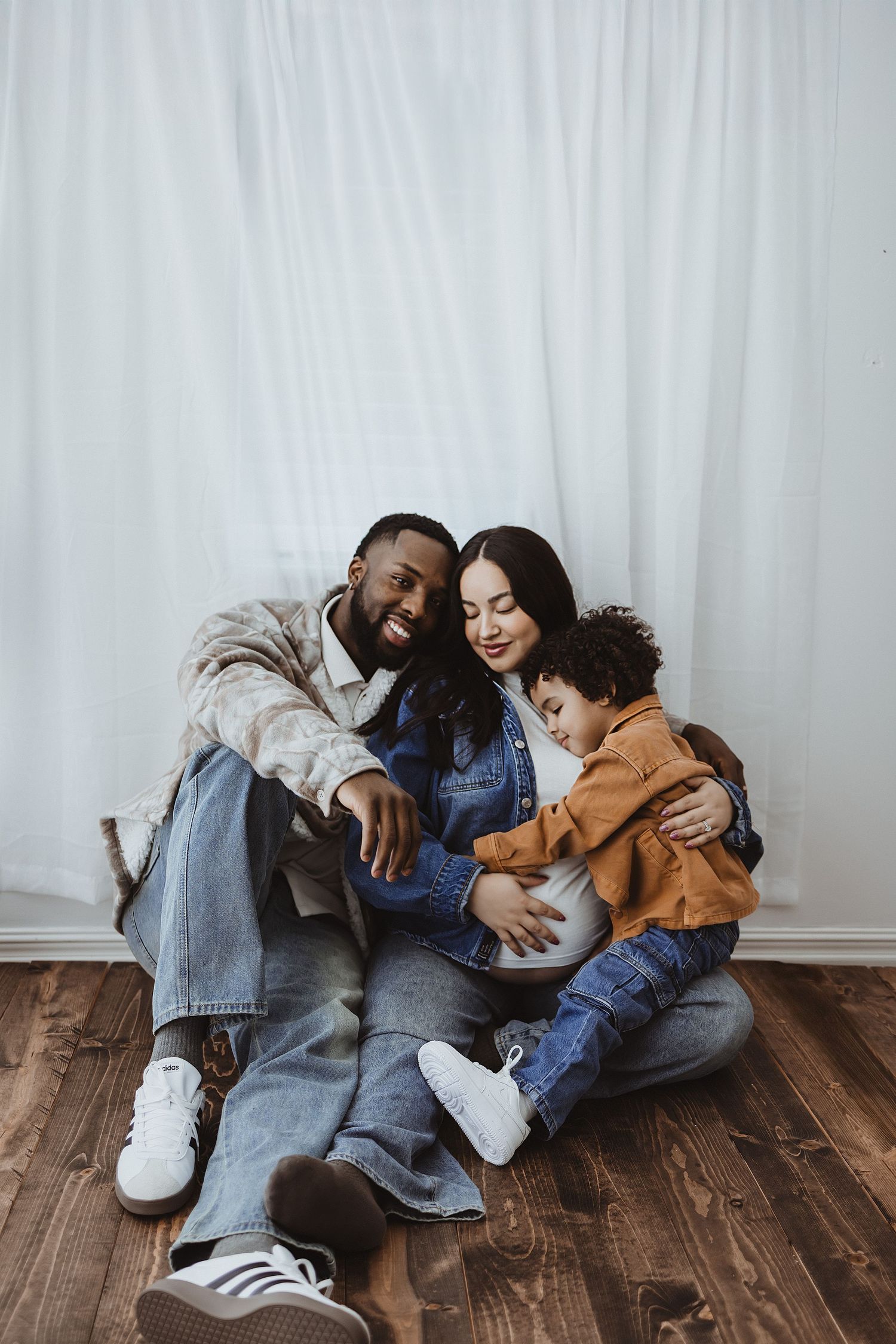 Happy interracial family sitting on wooden floor together with pregnant mother and young child.