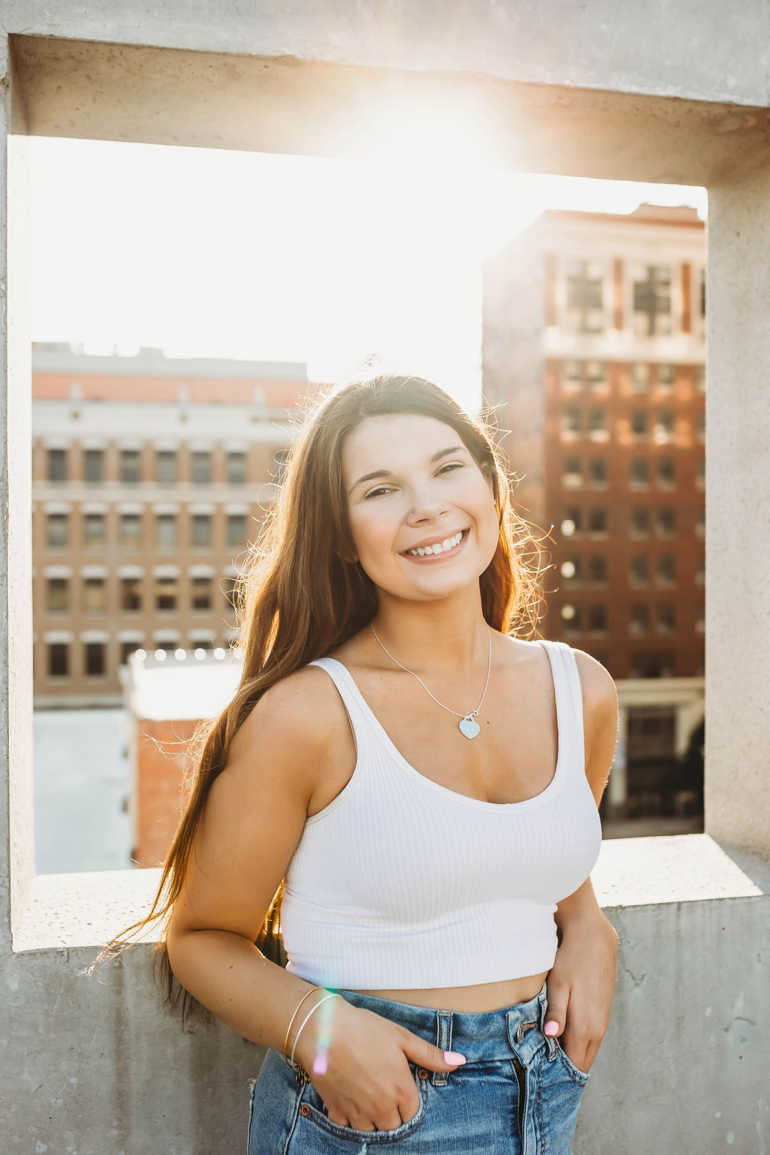 Smiling young woman in white crop top and jeans on rooftop with city buildings behind.