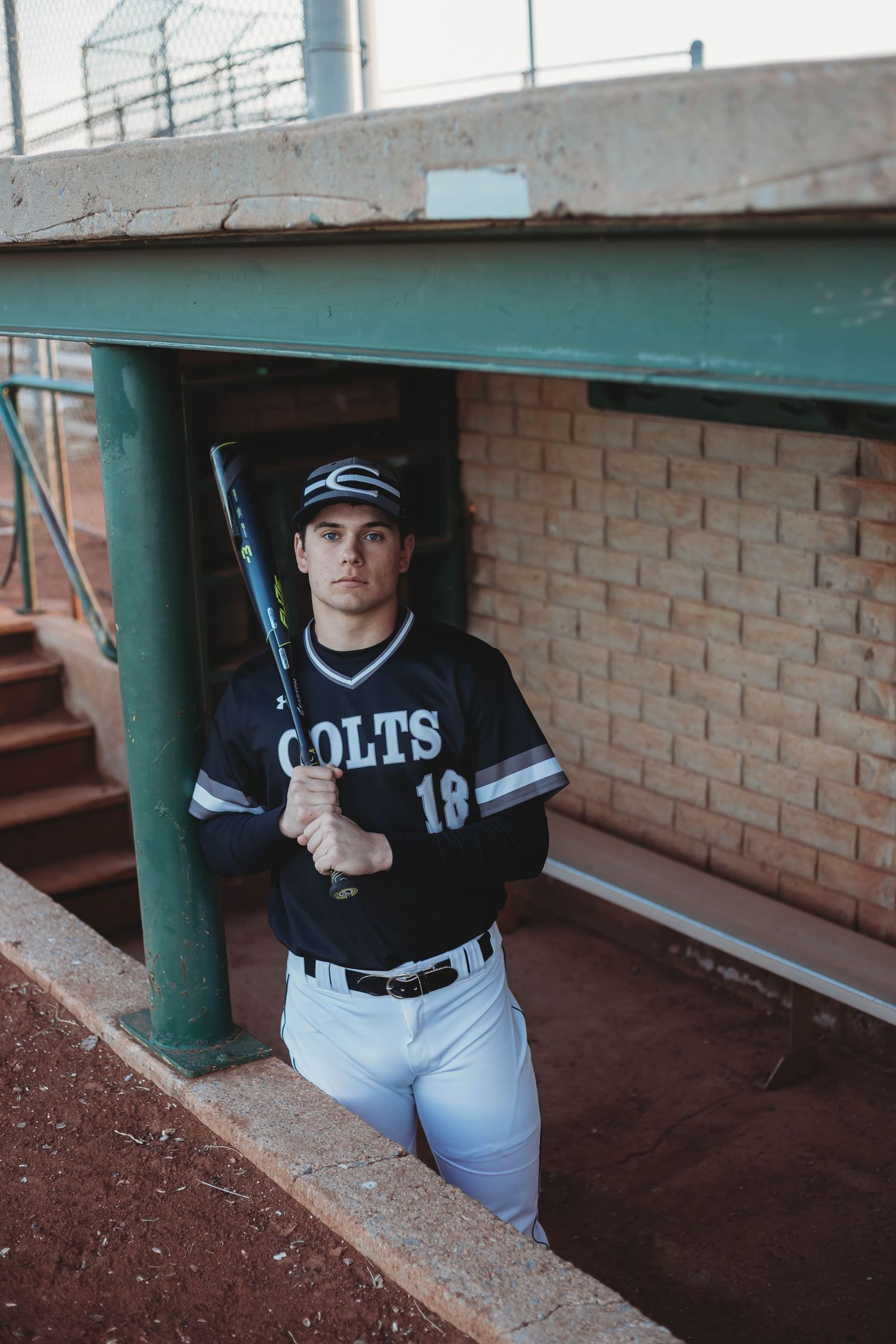 High school baseball player in Colts uniform number 18 holding a bat in a dugout.