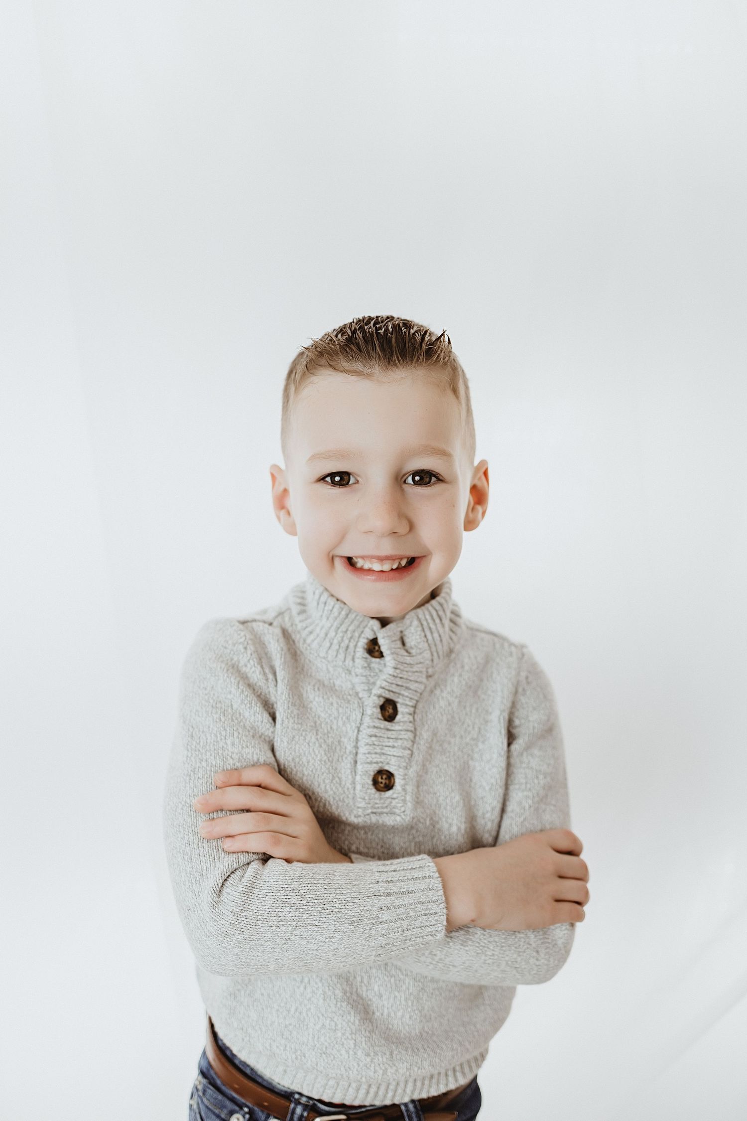 Happy young boy in gray sweater smiling with arms crossed against white background.